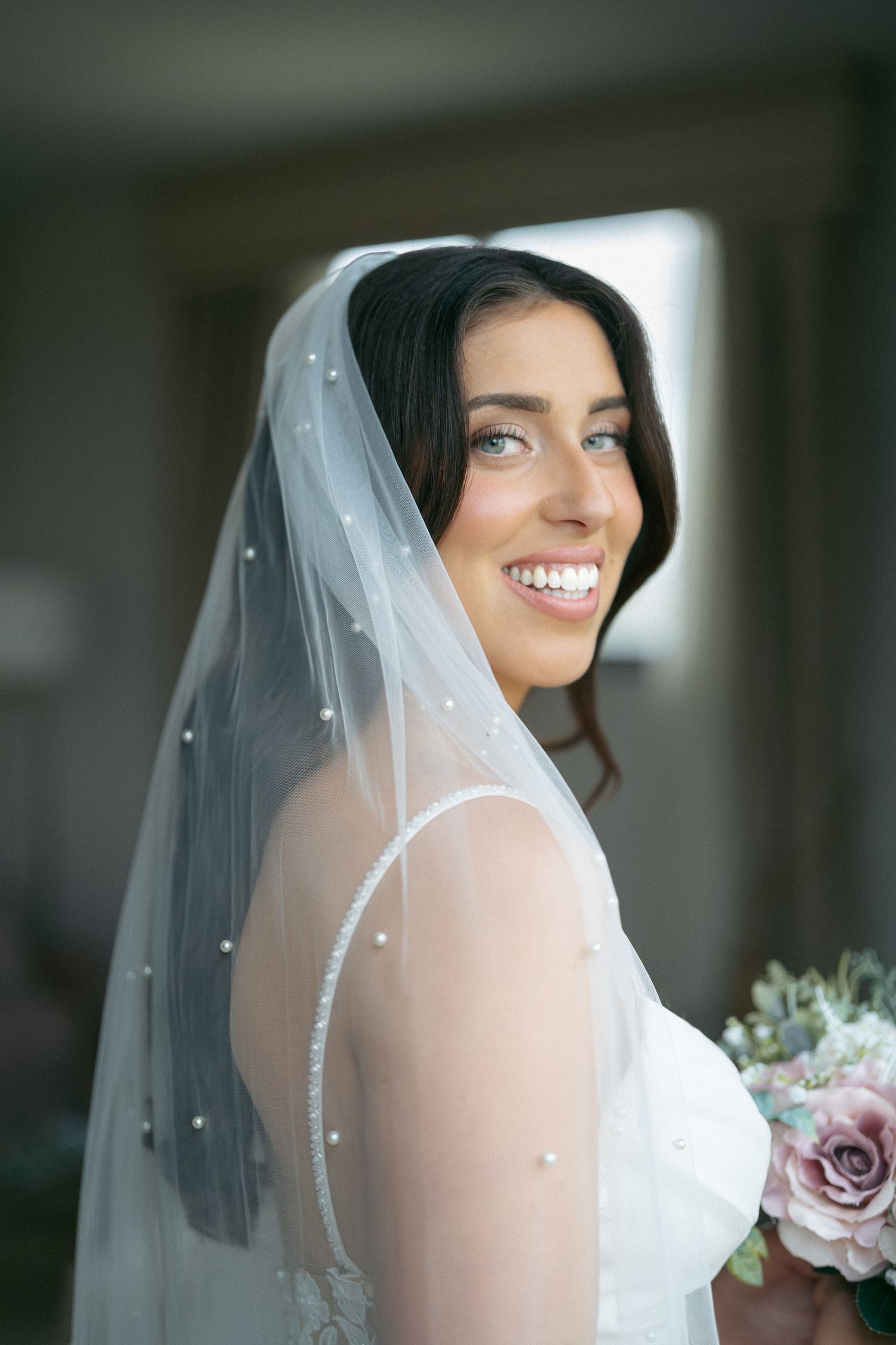 Joyful bride in a pearl-embellished veil smiles over her shoulder while holding a pastel rose bouquet.