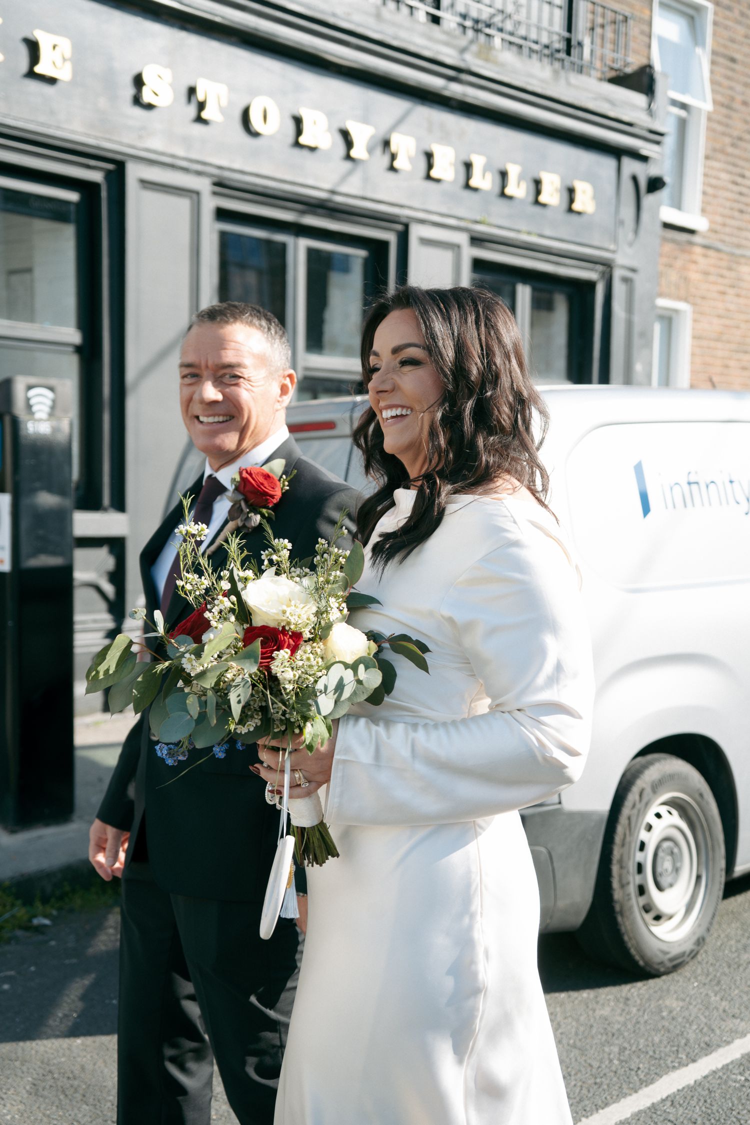 Laughing bride in a white dress and groom with a red rose boutonniere walk past a storefront holding a bouquet of white and red flowers.