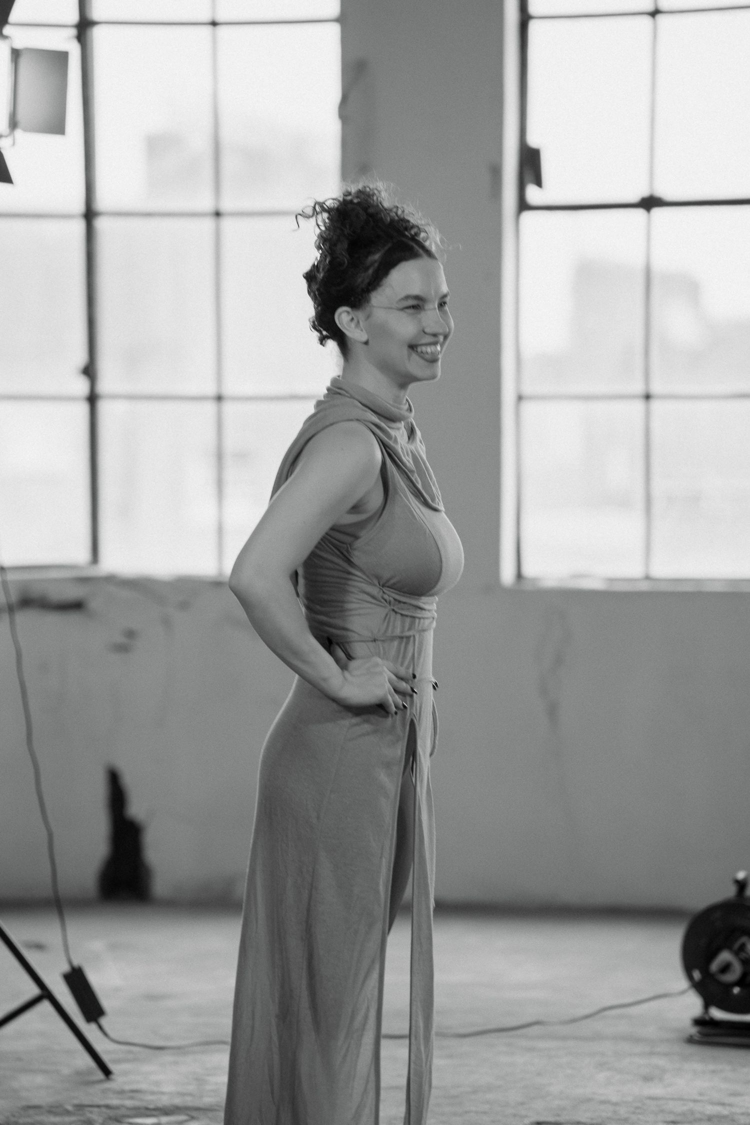 Smiling woman with curly updo in a sleeveless draped gown stands in a bright industrial studio with large windows in black and white.