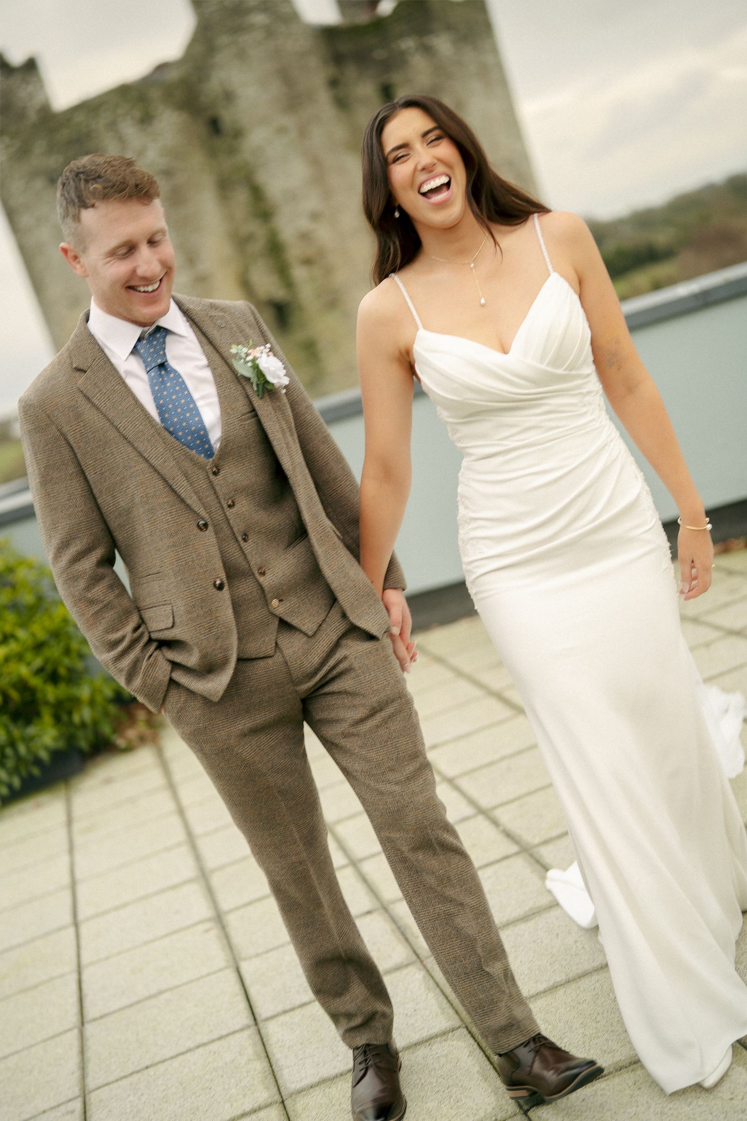 Laughing bride in a white dress walks hand in hand with a groom in a tweed suit on a rooftop terrace with a castle behind.