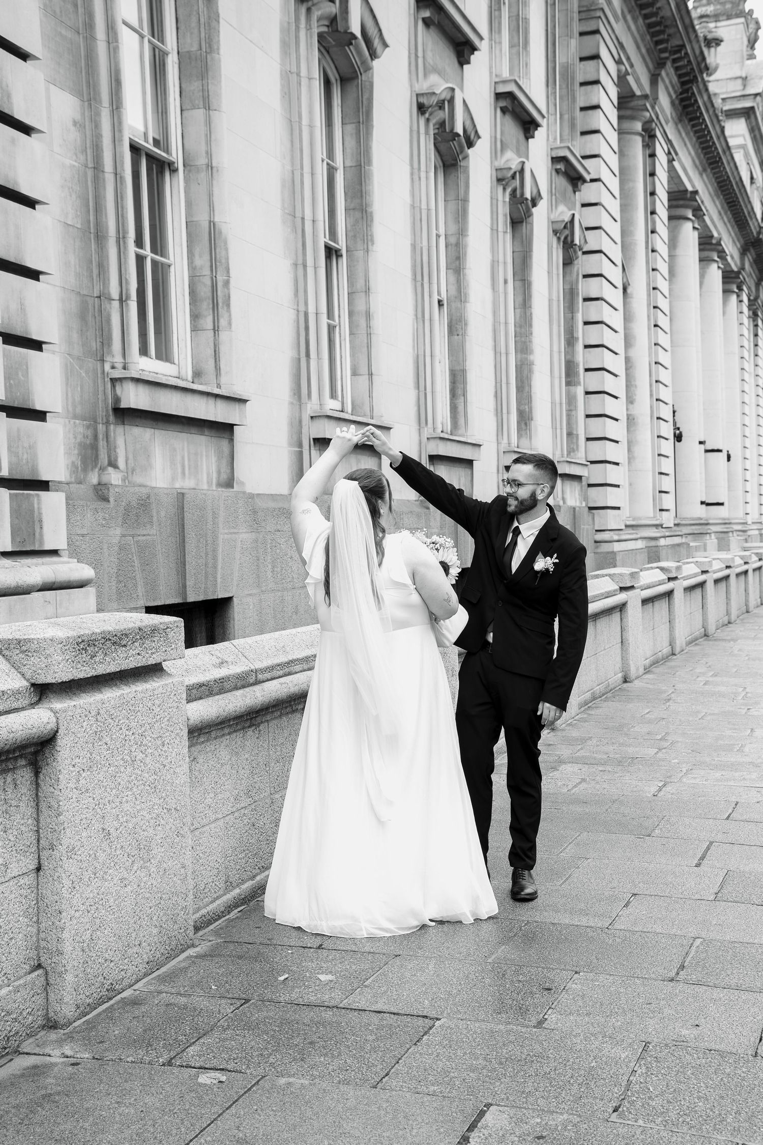 Groom twirls his bride in a long veil and white gown on a paved sidewalk beside classical stone columns in black and white.