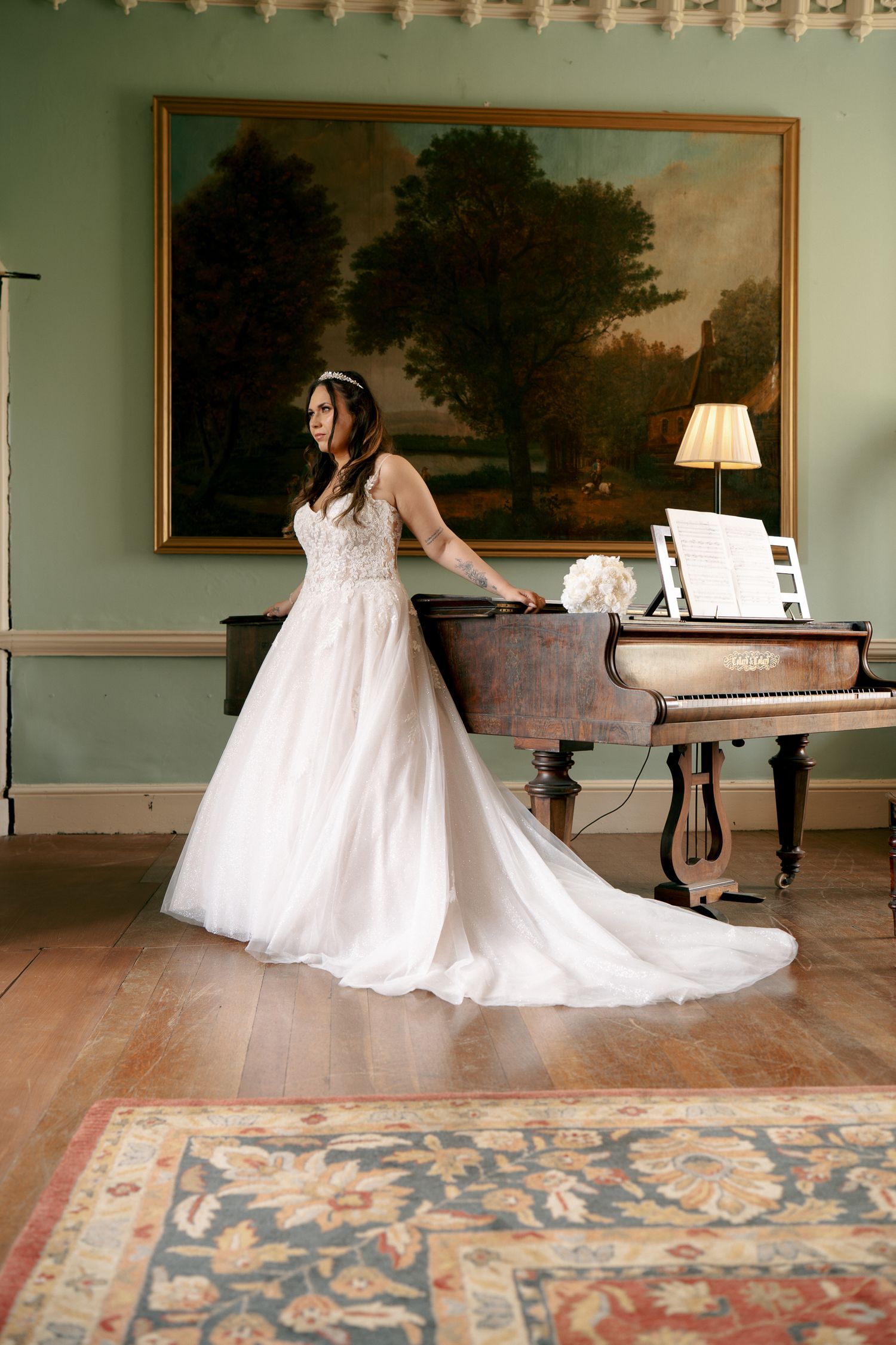 A bride in a long white gown with a flowing train stands beside a grand piano in an elegant room with a landscape painting.