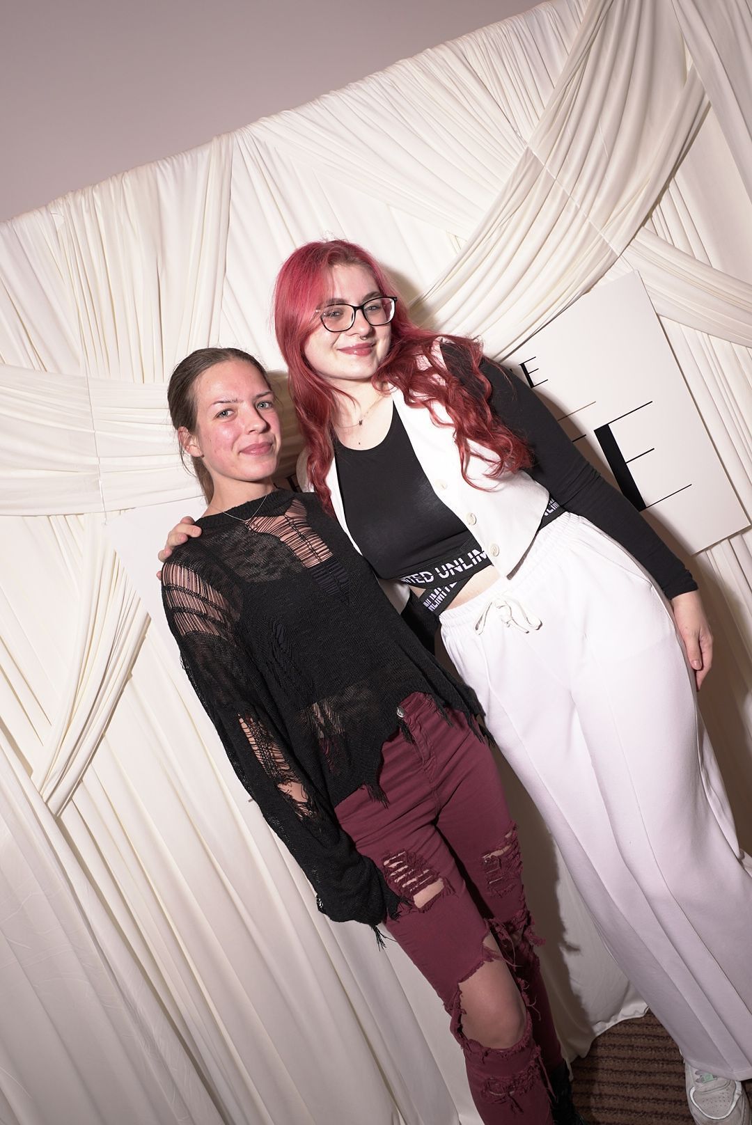 Two young women, one with red hair and glasses, pose together in front of a white draped event backdrop.