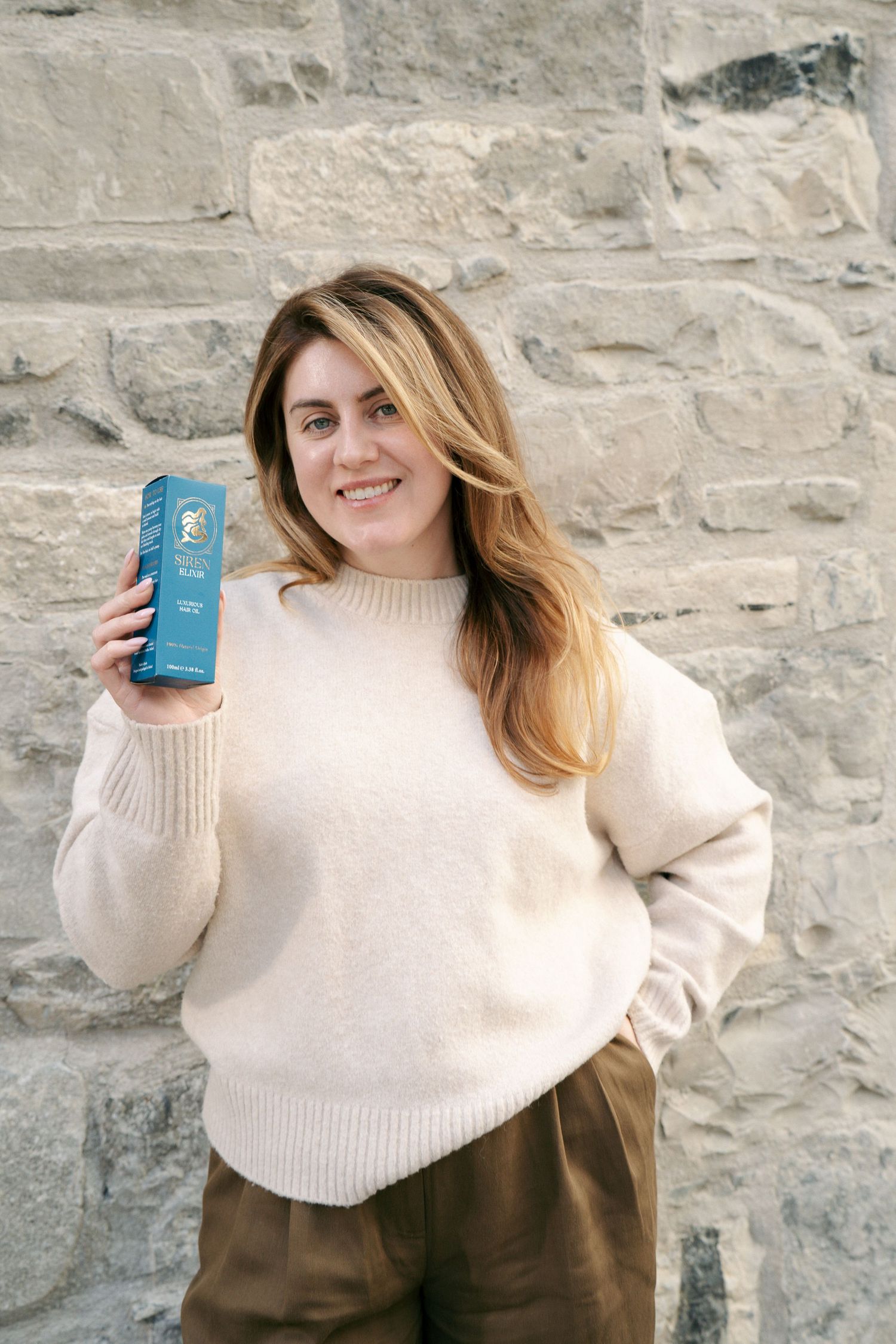 Smiling woman in a cream sweater holds a blue Siren Elixir hair oil box against a rustic stone wall.