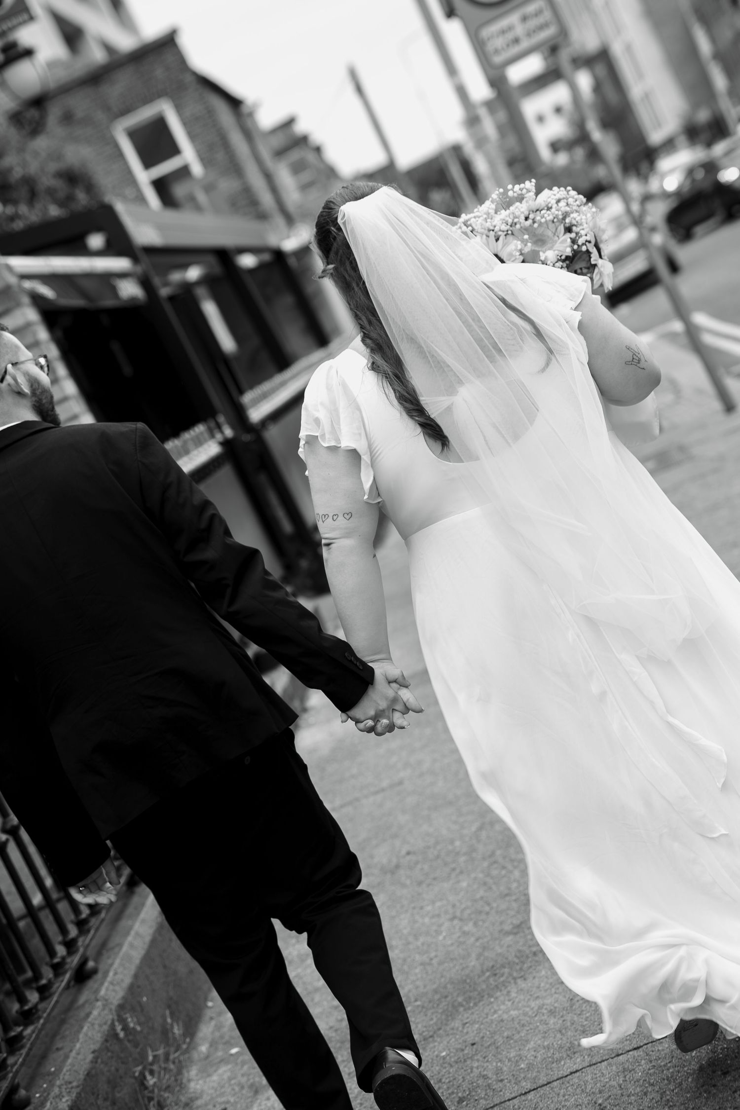 Bride and groom walk hand in hand down a city street, seen from behind, her veil and white gown flowing, black and white wedding photo.