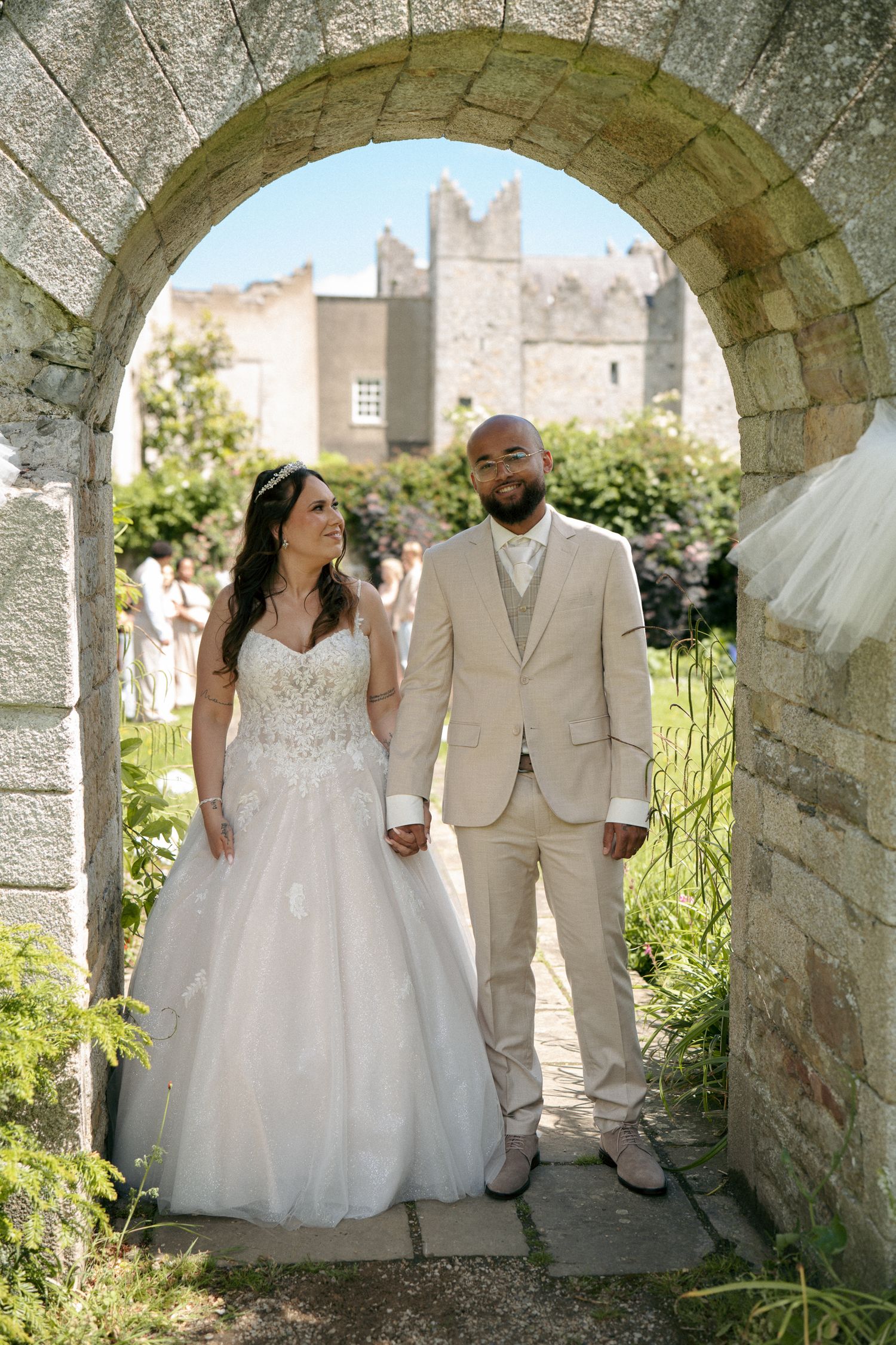A bride and groom hold hands under a stone archway in a garden, with a historic castle-like building behind them.