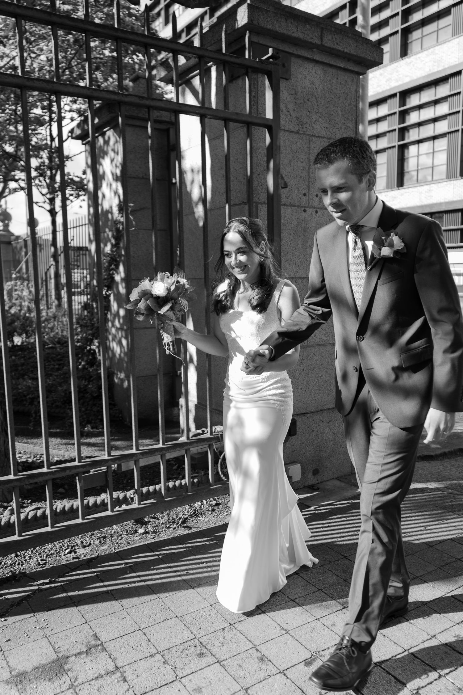 Black and white photo of a bride in a fitted gown holding a bouquet and walking hand in hand with her groom beside iron gates.