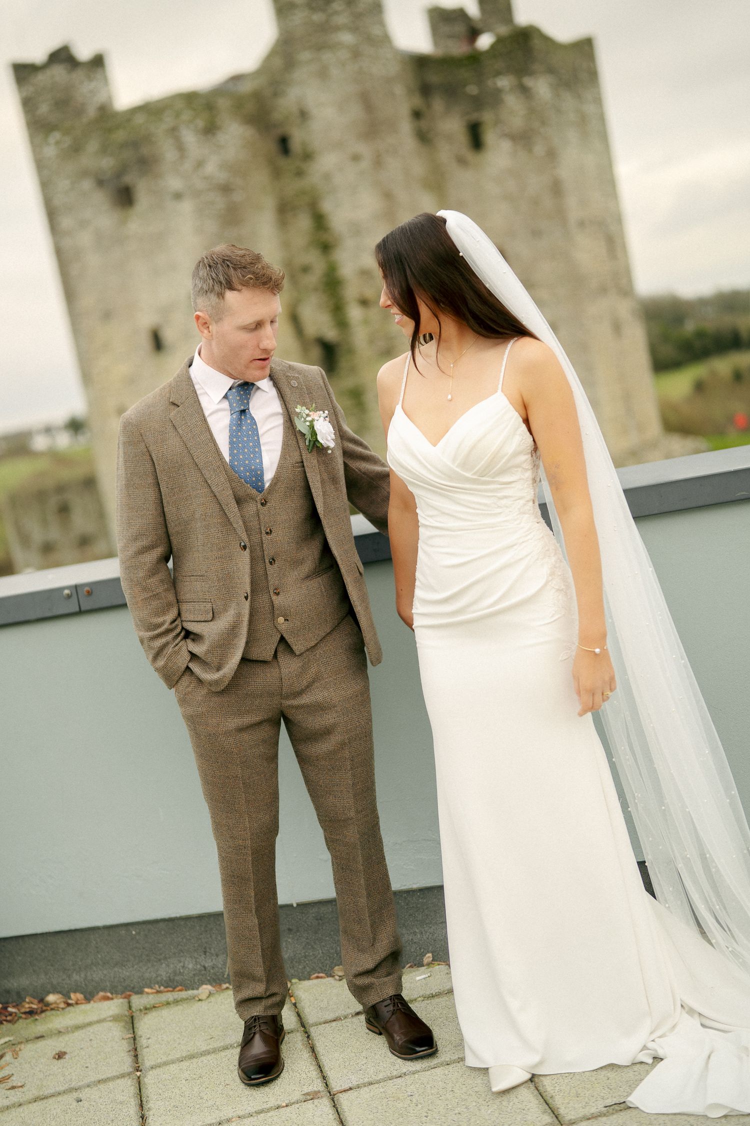 Groom in a brown tweed suit looks at his bride in a white gown and veil on a terrace in front of a stone castle.