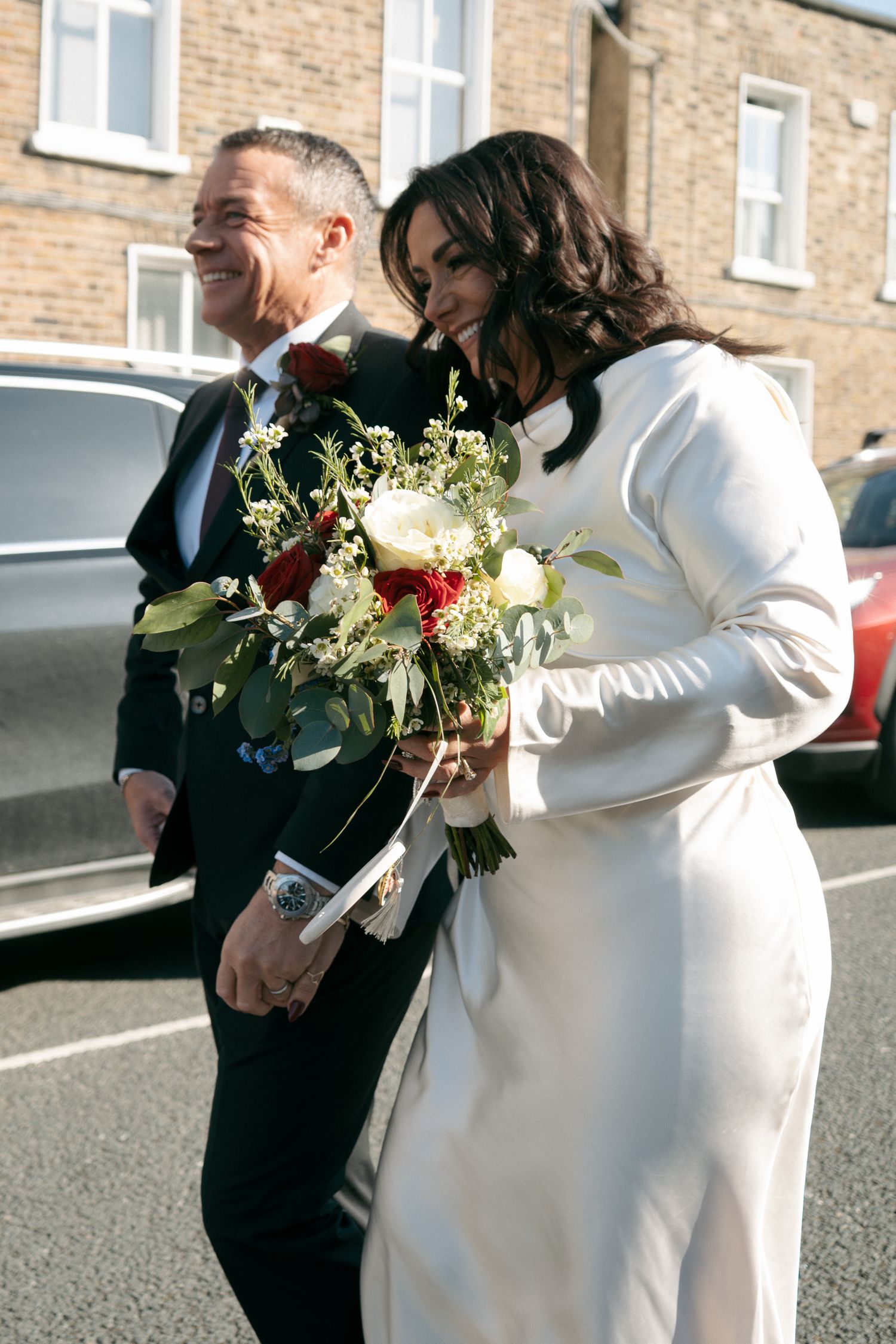 Bride in a white satin gown holds a red and white bouquet as she walks arm in arm with her smiling groom along a brick-lined street.