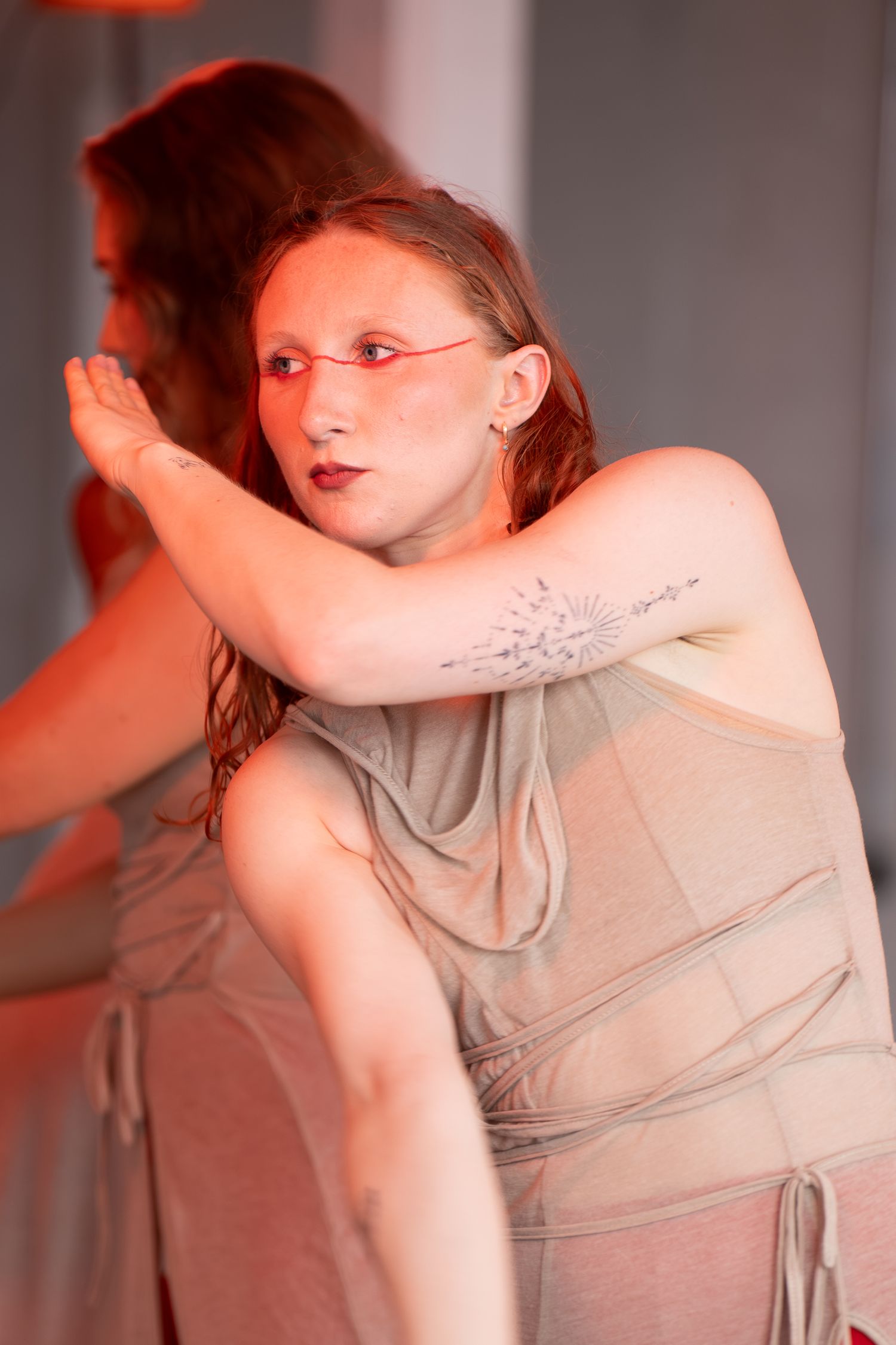 A dancer in a draped, tan wrap-style costume with a tattoo on her upper arm strikes an expressive pose with one hand raised near her face.