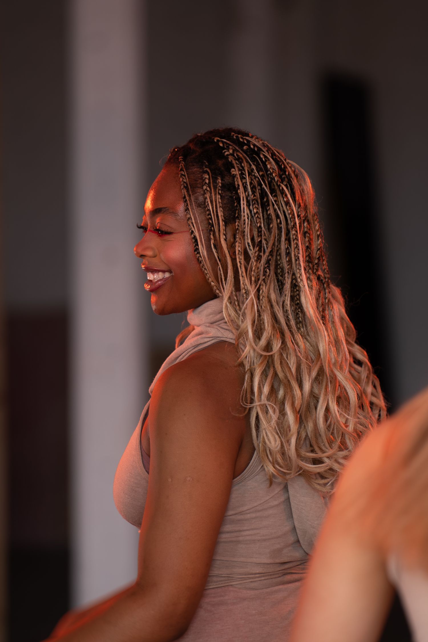 A smiling woman with long blonde-tipped braids is photographed in profile in warm orange-toned lighting.
