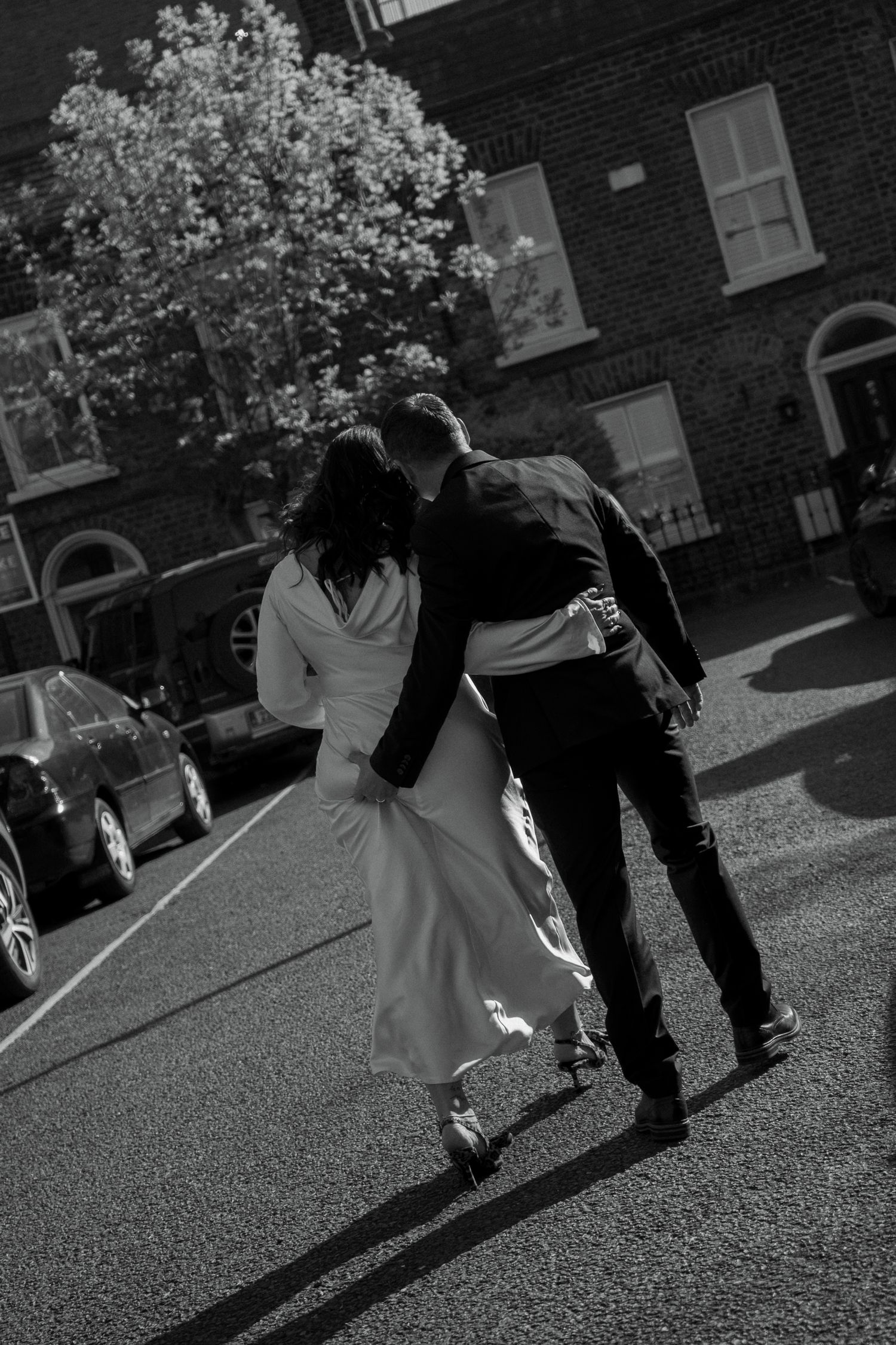 Black and white photo of a couple walking away down a city street, the groom's arm around the bride in her flowing satin wedding gown.