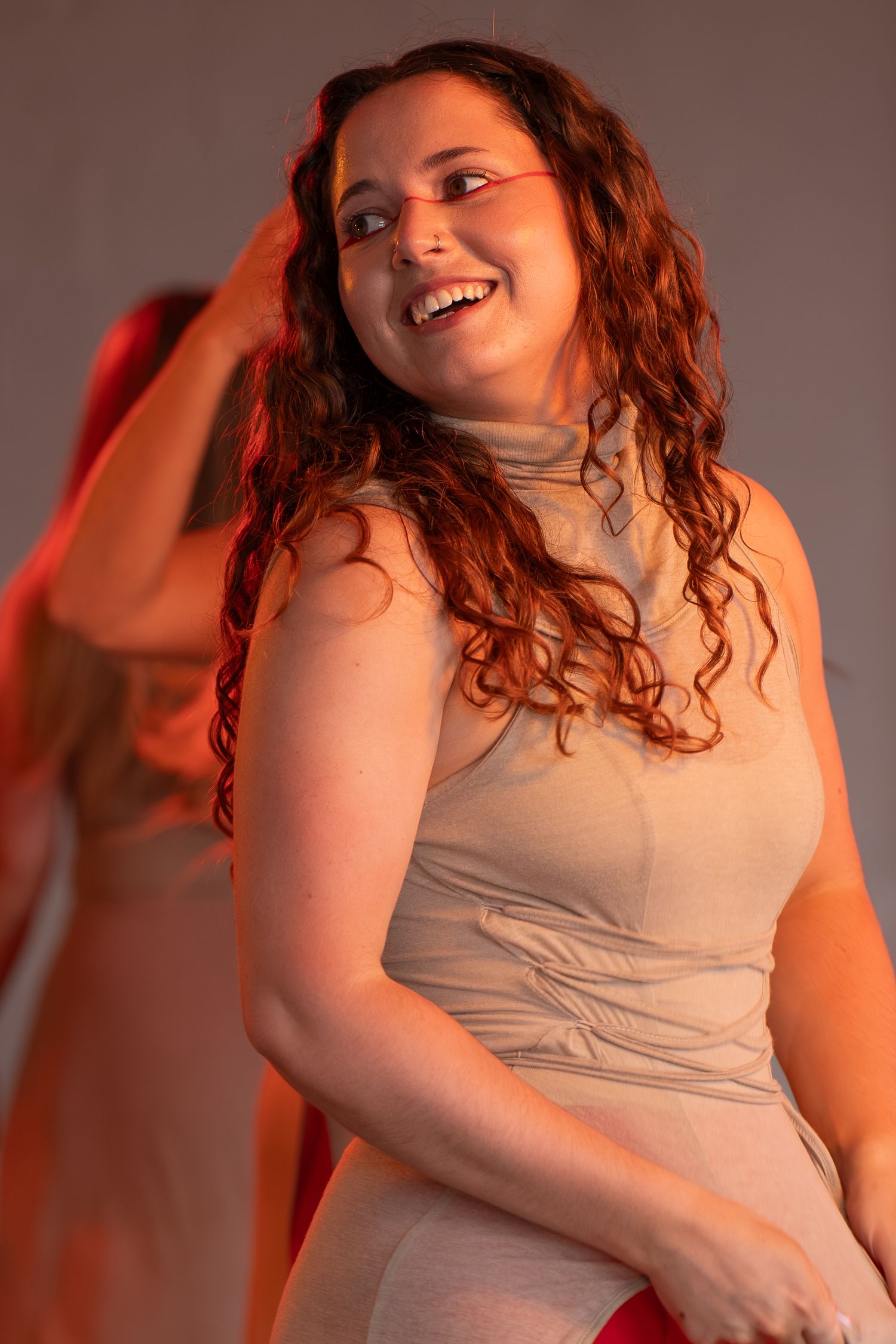 Smiling woman with long curly auburn hair in a beige sleeveless turtleneck dress glances over her shoulder while performing on stage.