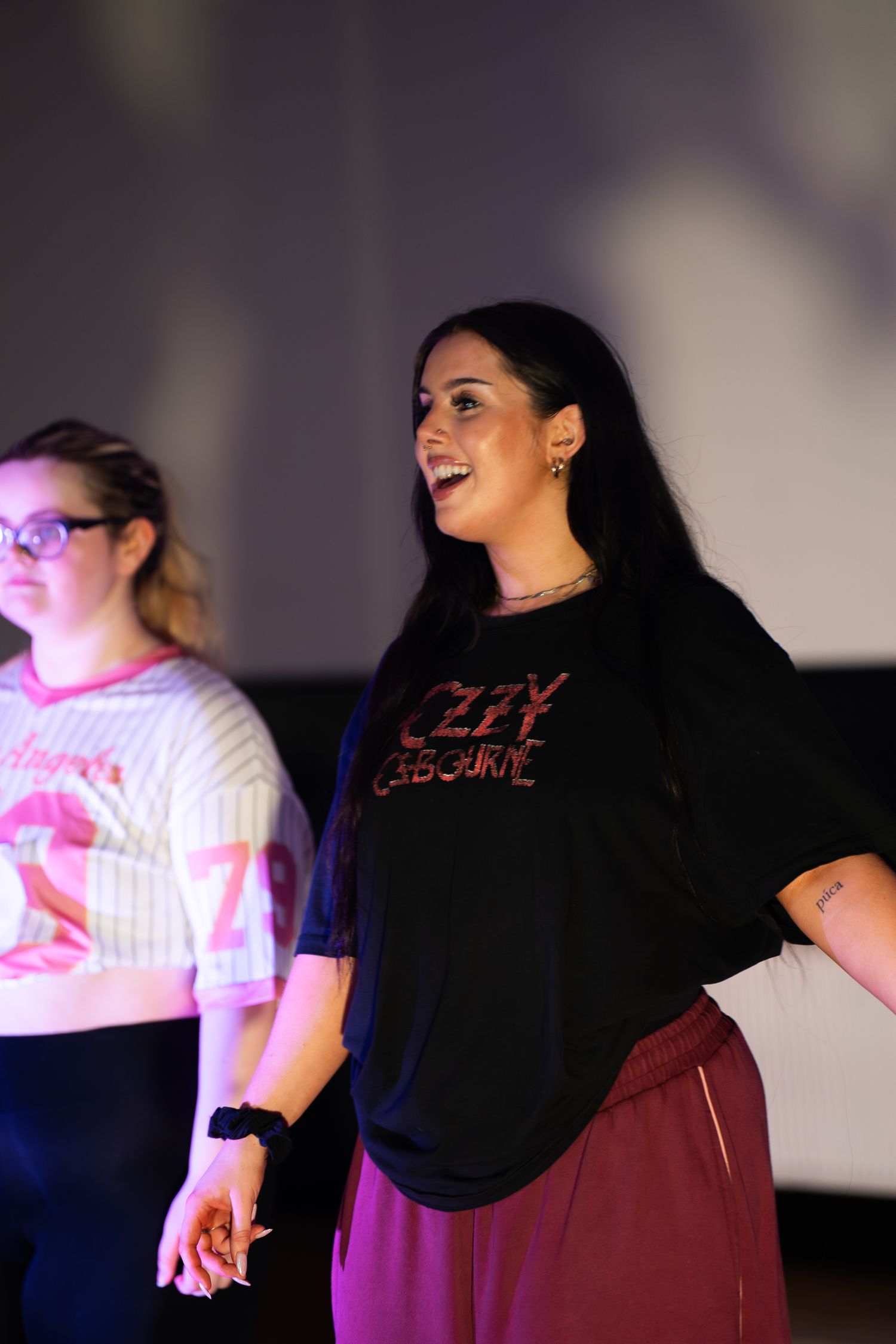 Smiling woman with long dark hair in an Ozzy Osbourne t-shirt and maroon pants dances on stage beside a girl in glasses and a pink top.