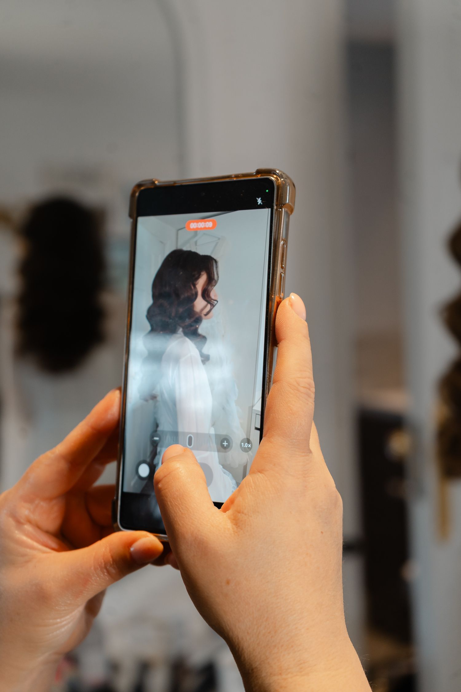 A hand holds a smartphone showing a camera view of a woman with dark curled hair in a white dress at a bridal hair styling session.