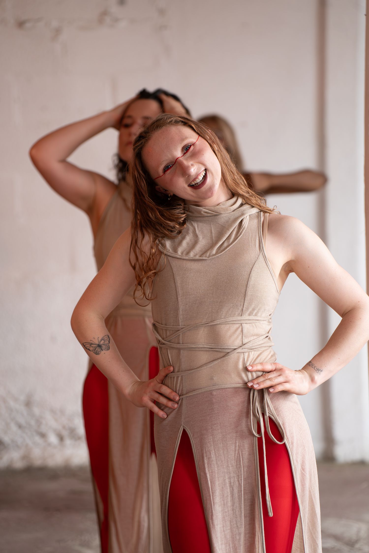 Smiling dancer in a beige sleeveless top and red pants poses with hands on hips as two dancers move behind her in a bright studio.
