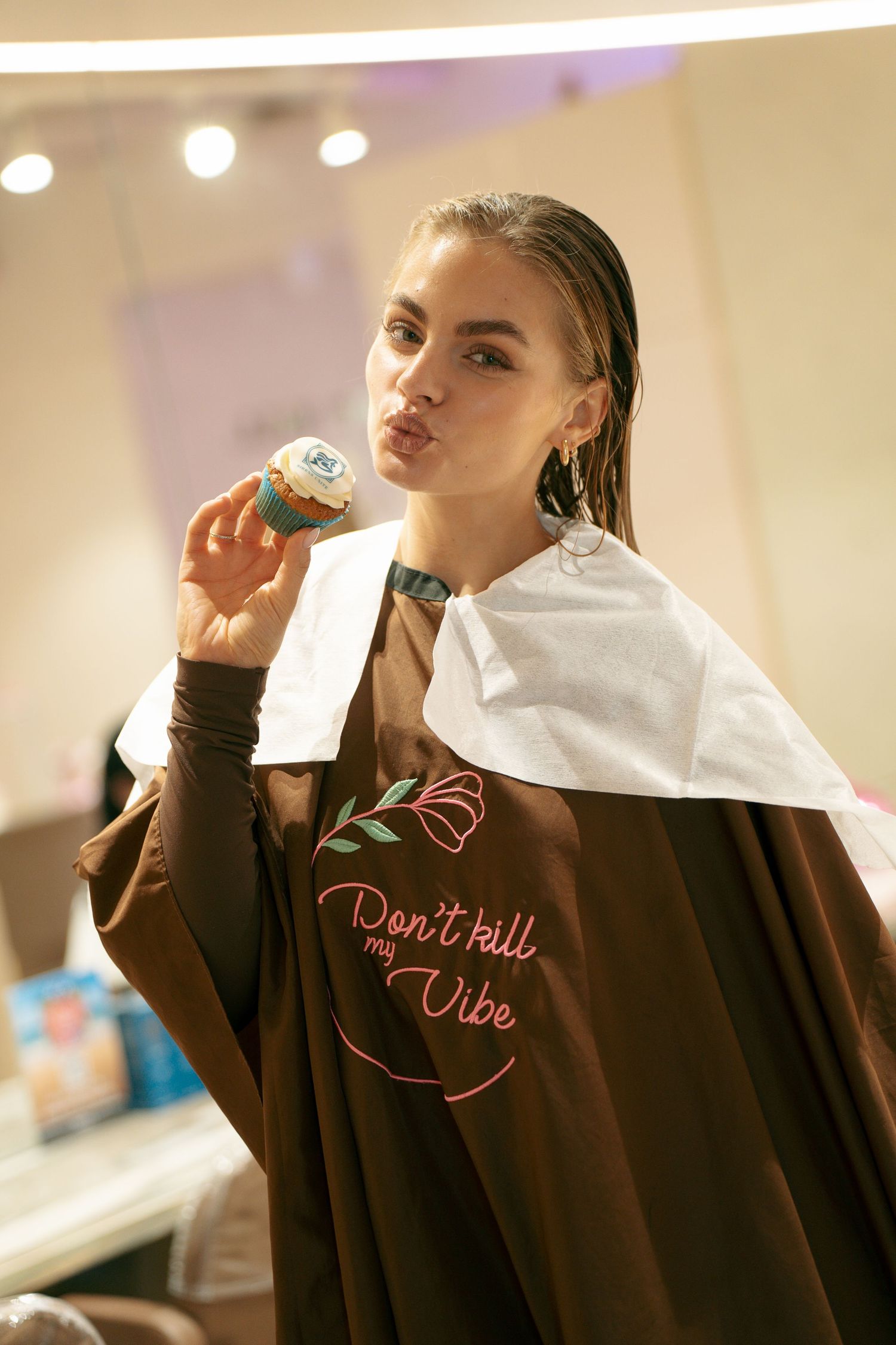 Woman in a brown salon cape reading Don't kill my vibe playfully holds a logo cupcake with pursed lips in a bright salon.
