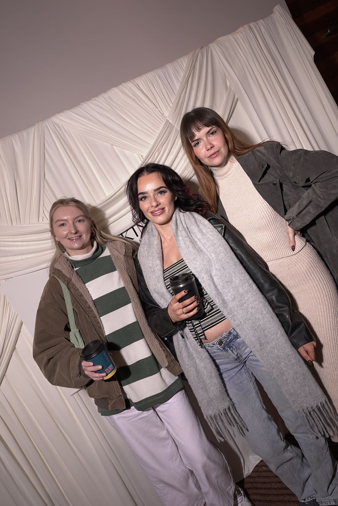 Three women in winter coats and scarves, holding coffee cups, pose together in front of a white draped fabric backdrop at an event.