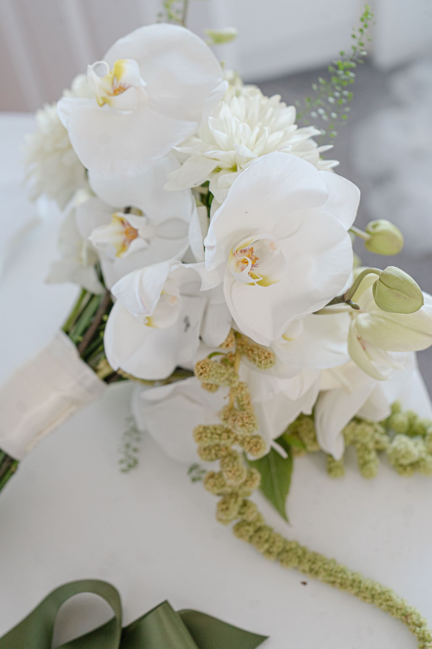 Close-up of a white orchid and dahlia bridal bouquet with green amaranthus accents, wrapped in cream ribbon and resting on a table.