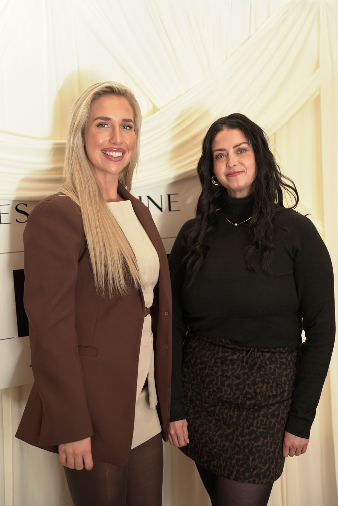 Two smiling women in stylish outfits, one in a brown blazer and one in black with a leopard skirt, pose in front of draped fabric.