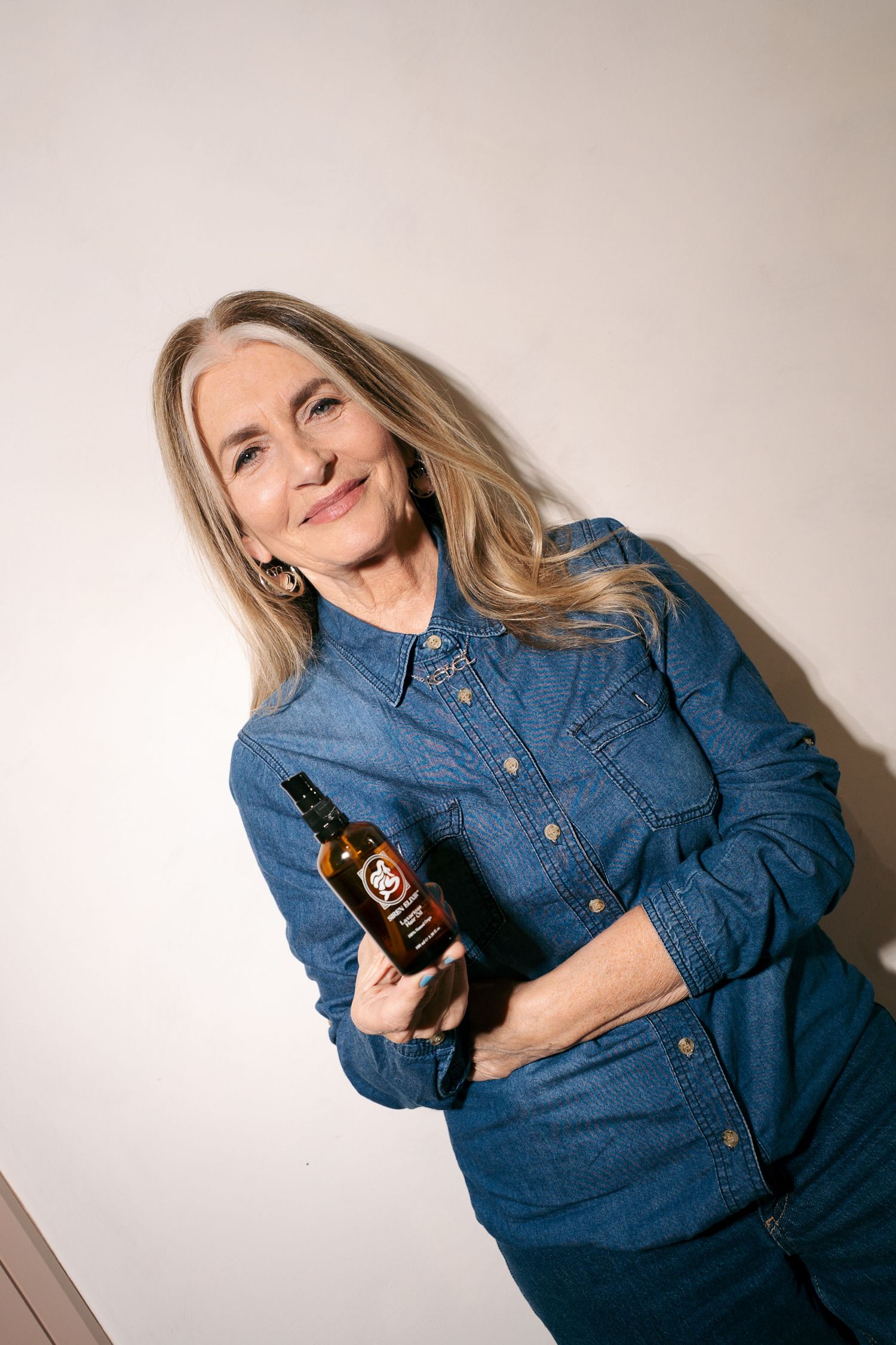 Smiling woman with long blonde hair in a denim shirt holds a small amber bottle of hair oil against a plain white wall.
