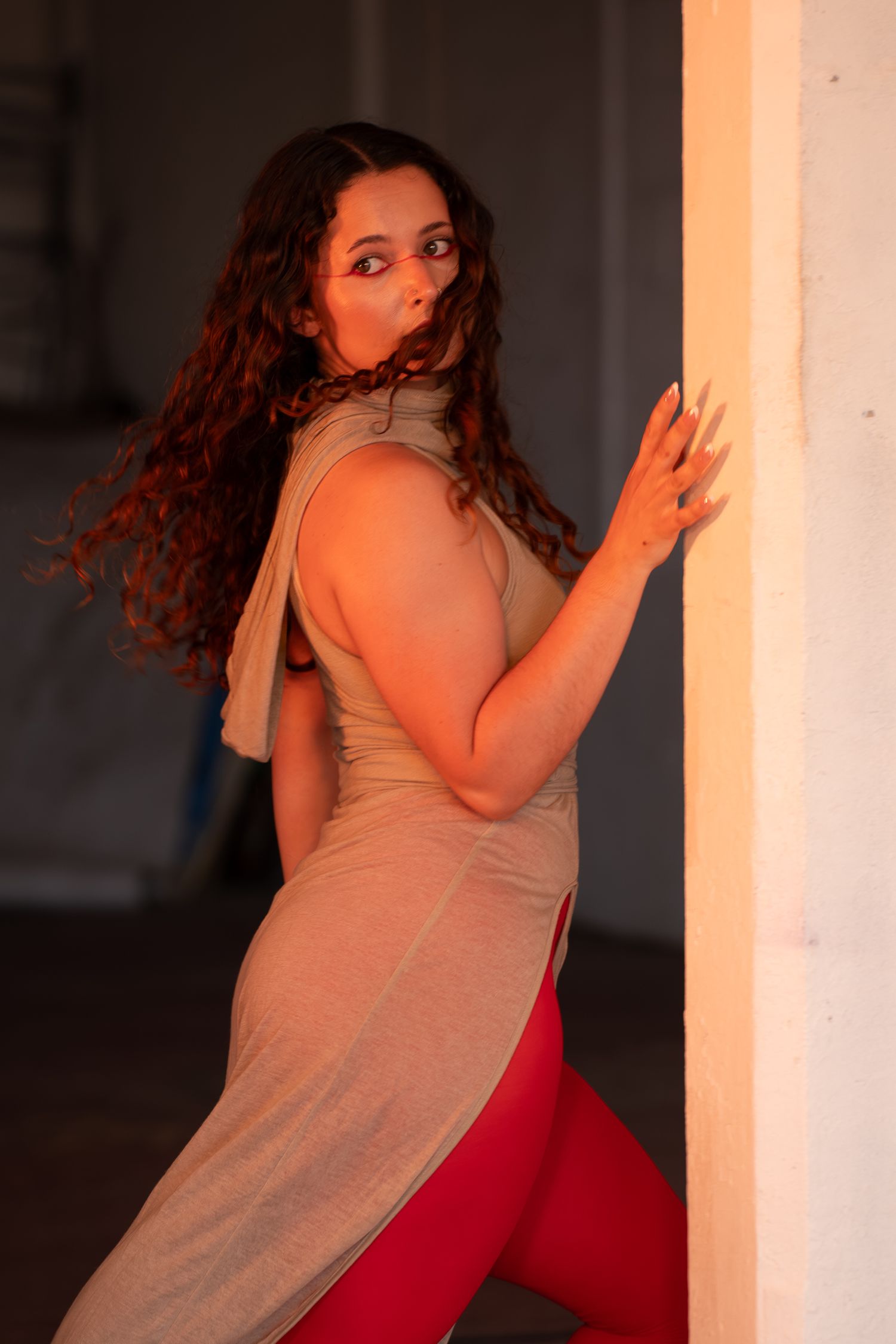 Woman with long curly hair in a tan sleeveless tunic and red leggings glances over her shoulder while leaning against a white wall.