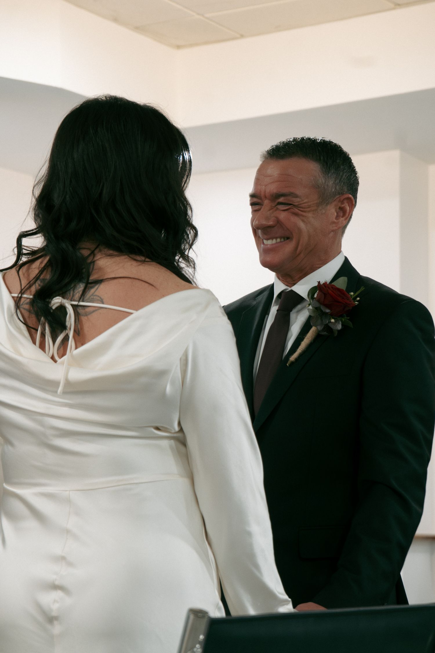 Groom in a black suit with a red boutonniere smiles warmly at his bride during a wedding ceremony.