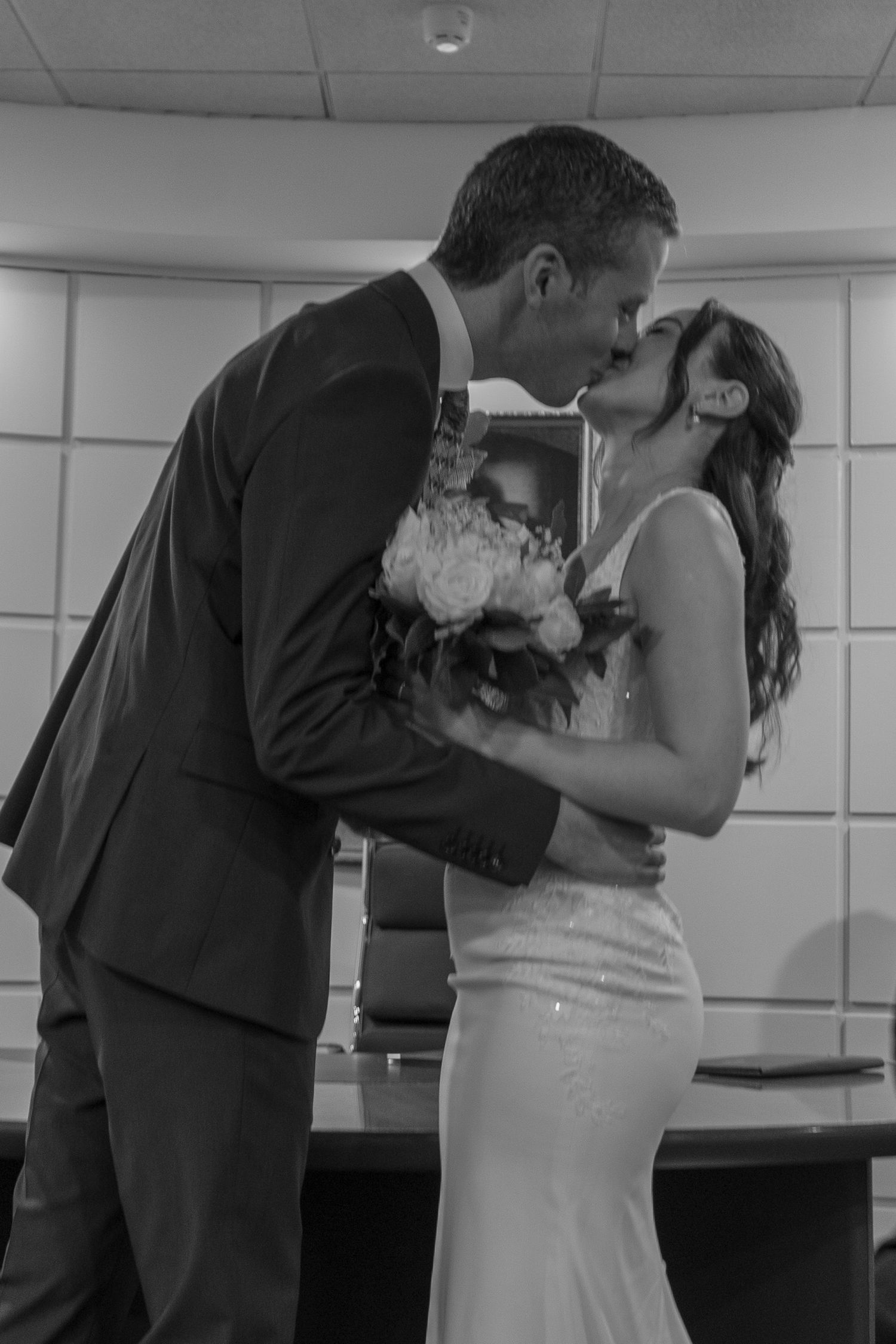 Black and white photo of a bride and groom sharing a kiss during their wedding ceremony, with the bride holding a bouquet.