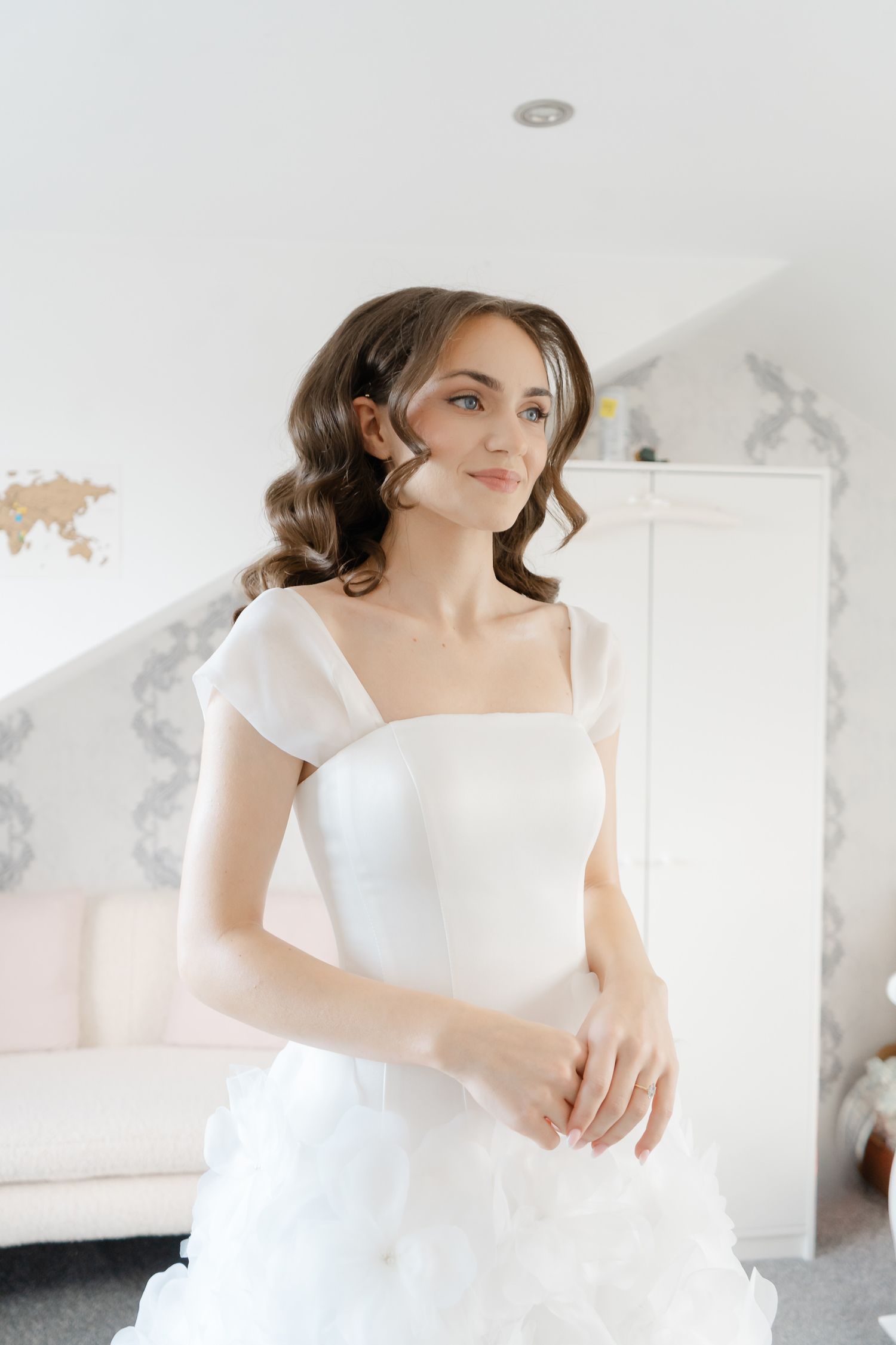 Bride in an off-the-shoulder white gown with a ruffled skirt gazing to the side in a bright, softly lit bedroom on her wedding day.