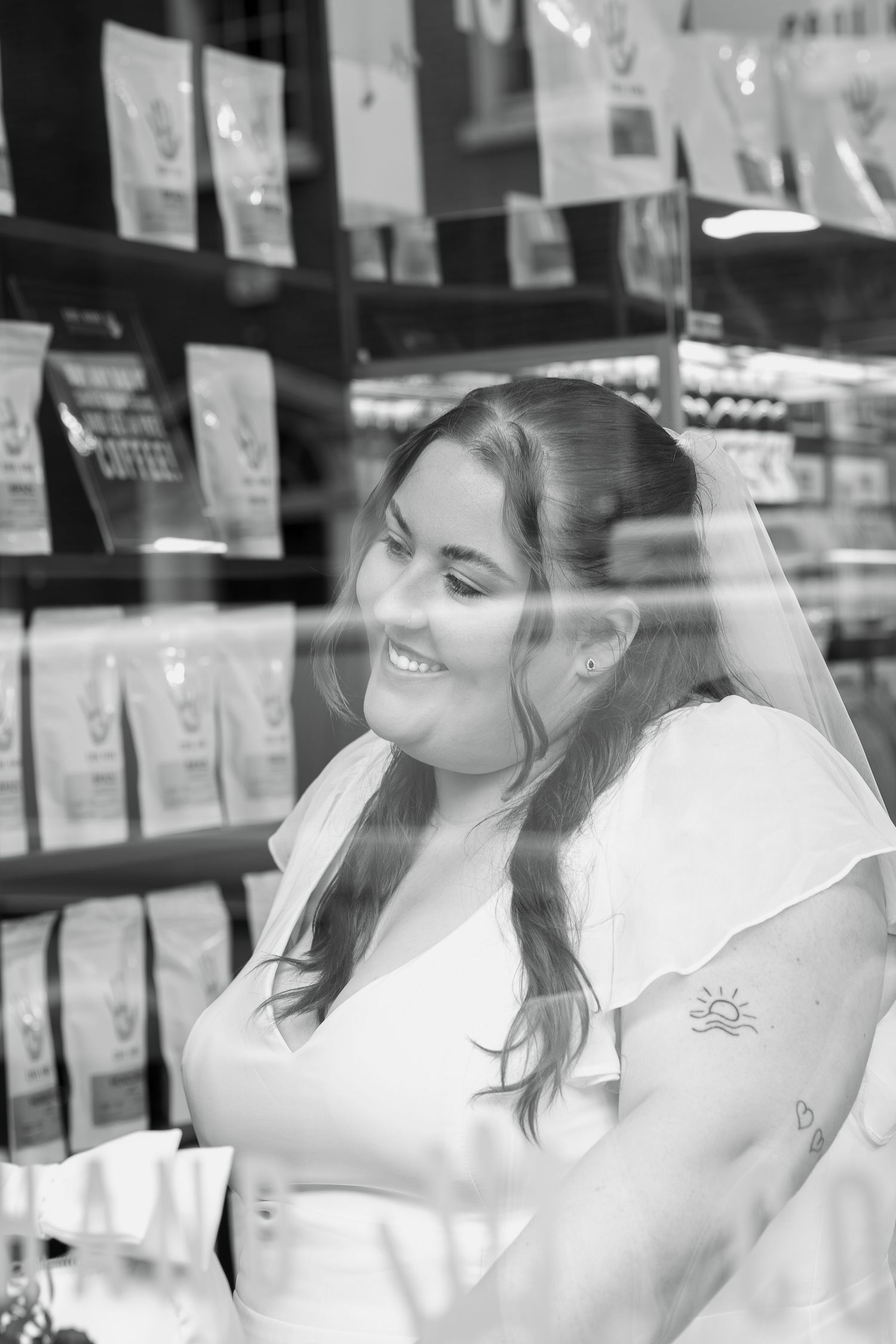 A smiling bride in a white dress and veil is seen through a shop window with coffee bags on the shelves behind her.
