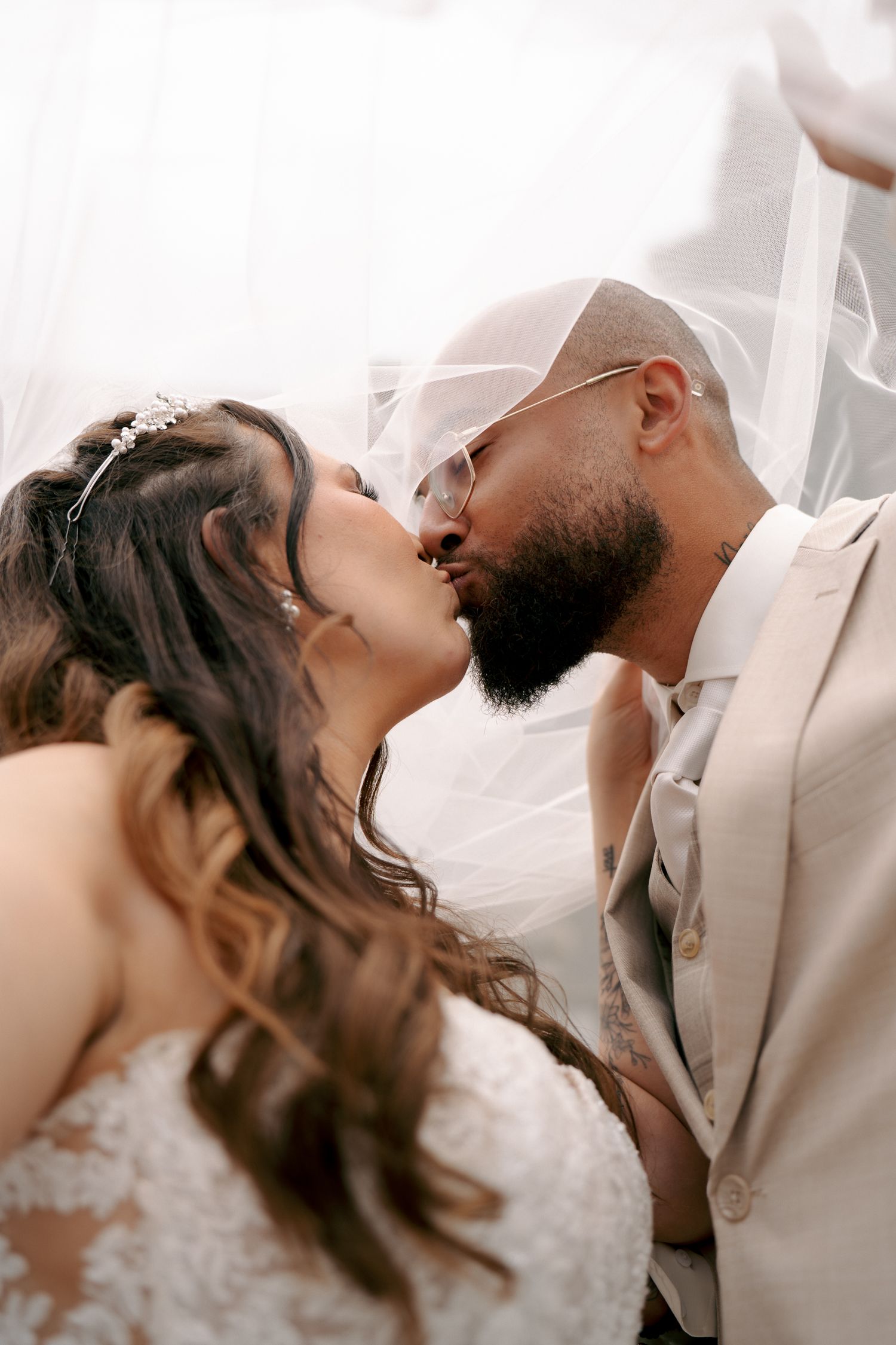 Groom with a beard and glasses kisses his bride in a lace gown and tiara as a sheer white veil drapes around them.