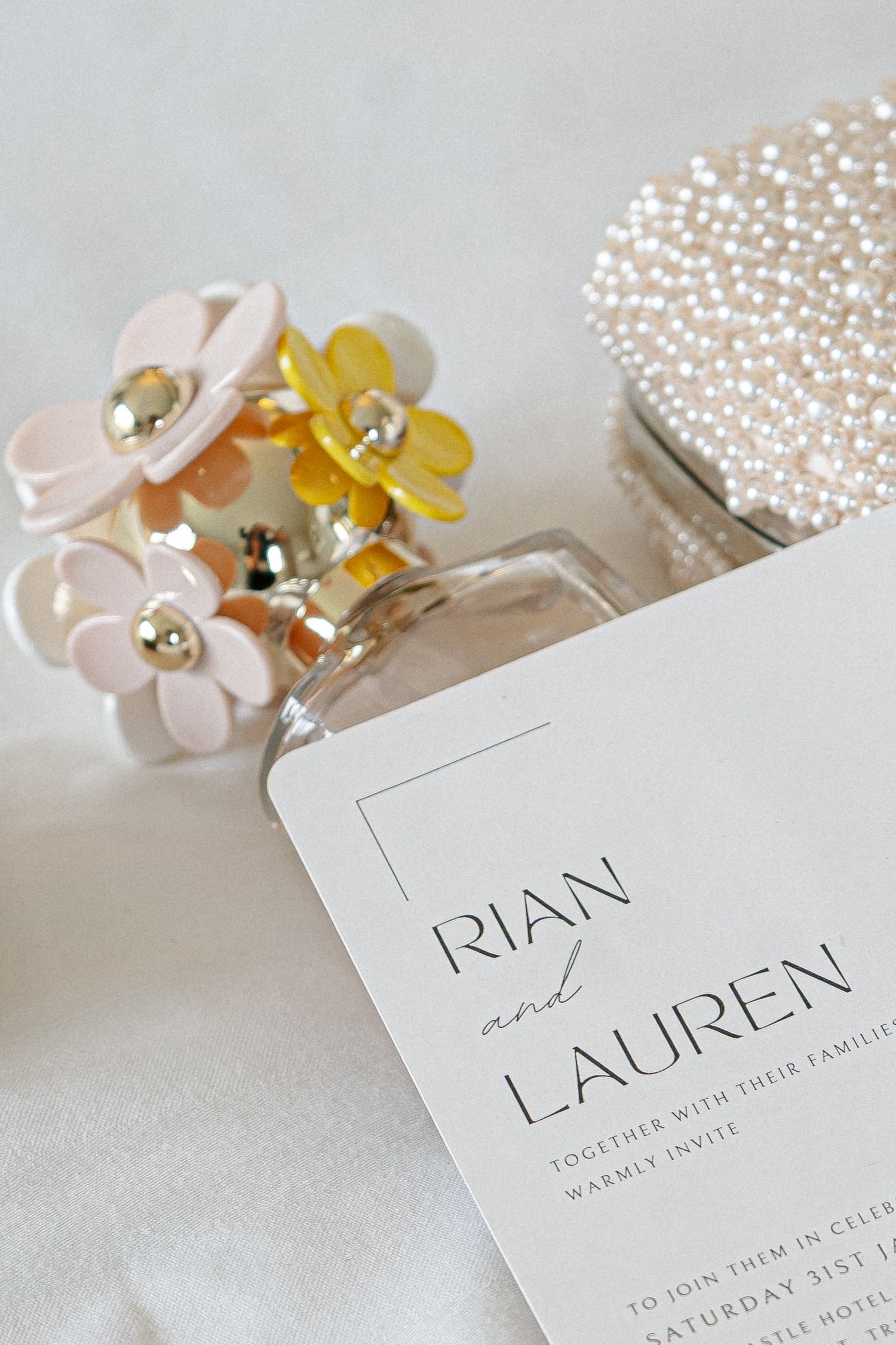 Wedding invitation for Rian and Lauren beside a pearl clutch and a perfume bottle with pastel flower decorations.