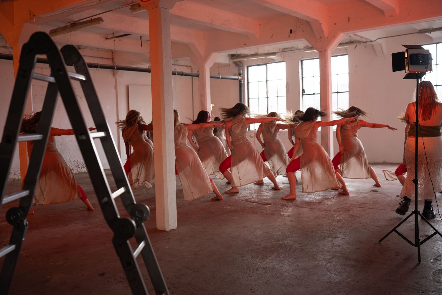 A group of dancers in flowing pale dresses perform in a line inside a large industrial studio while a woman stands beside a stage light.