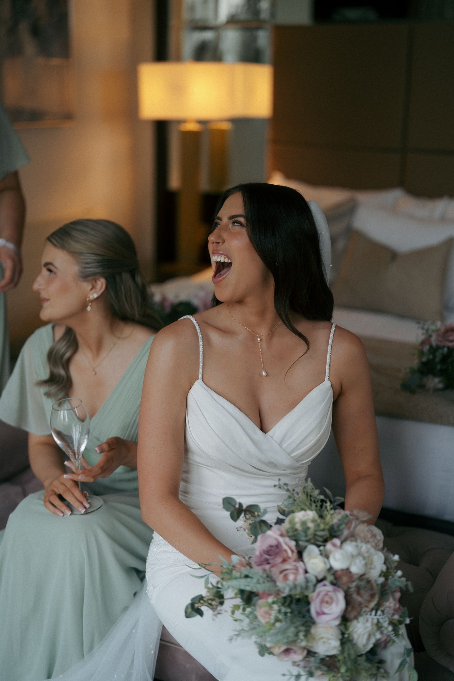 Laughing bride in a white dress holds a pink and white bouquet beside a bridesmaid in a sage green dress holding a glass.