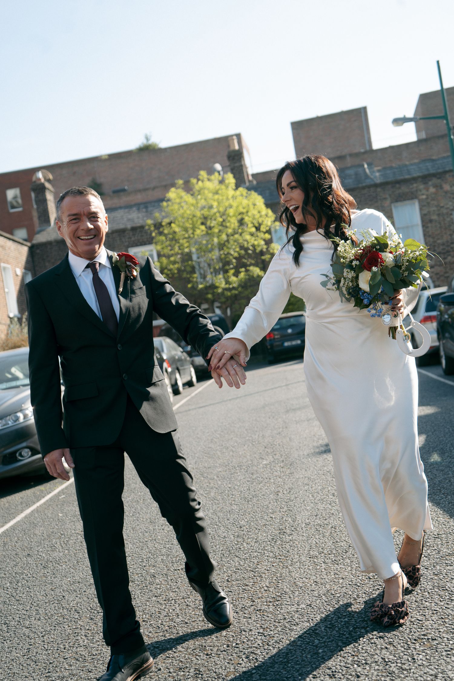 Happy newlywed couple holds hands and walks down a city street, the bride carrying a bouquet and wearing a sleek white wedding dress.