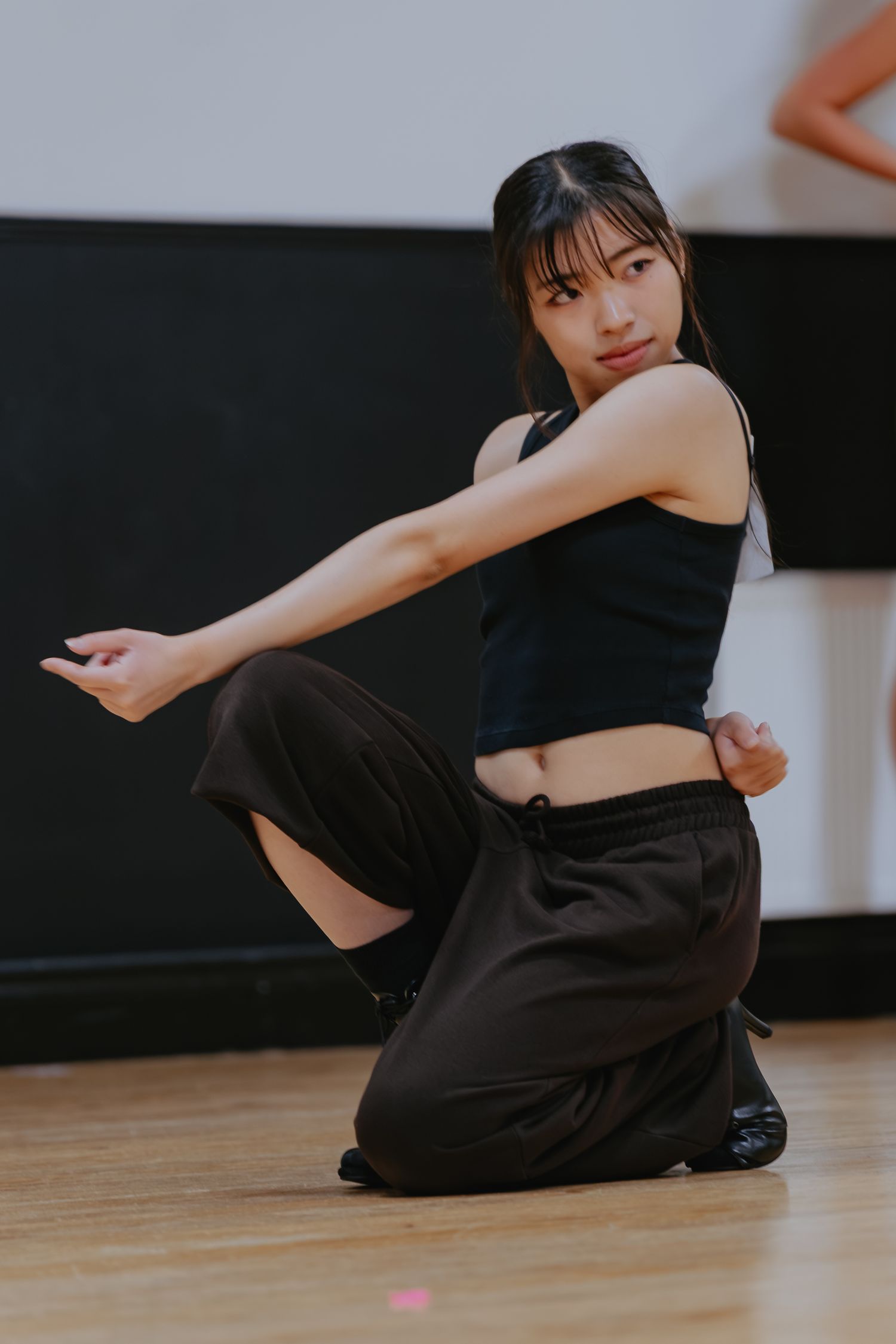 Dancer in a black tank top and brown cargo pants kneels on a wood studio floor, arms extended in a mid-choreography pose.