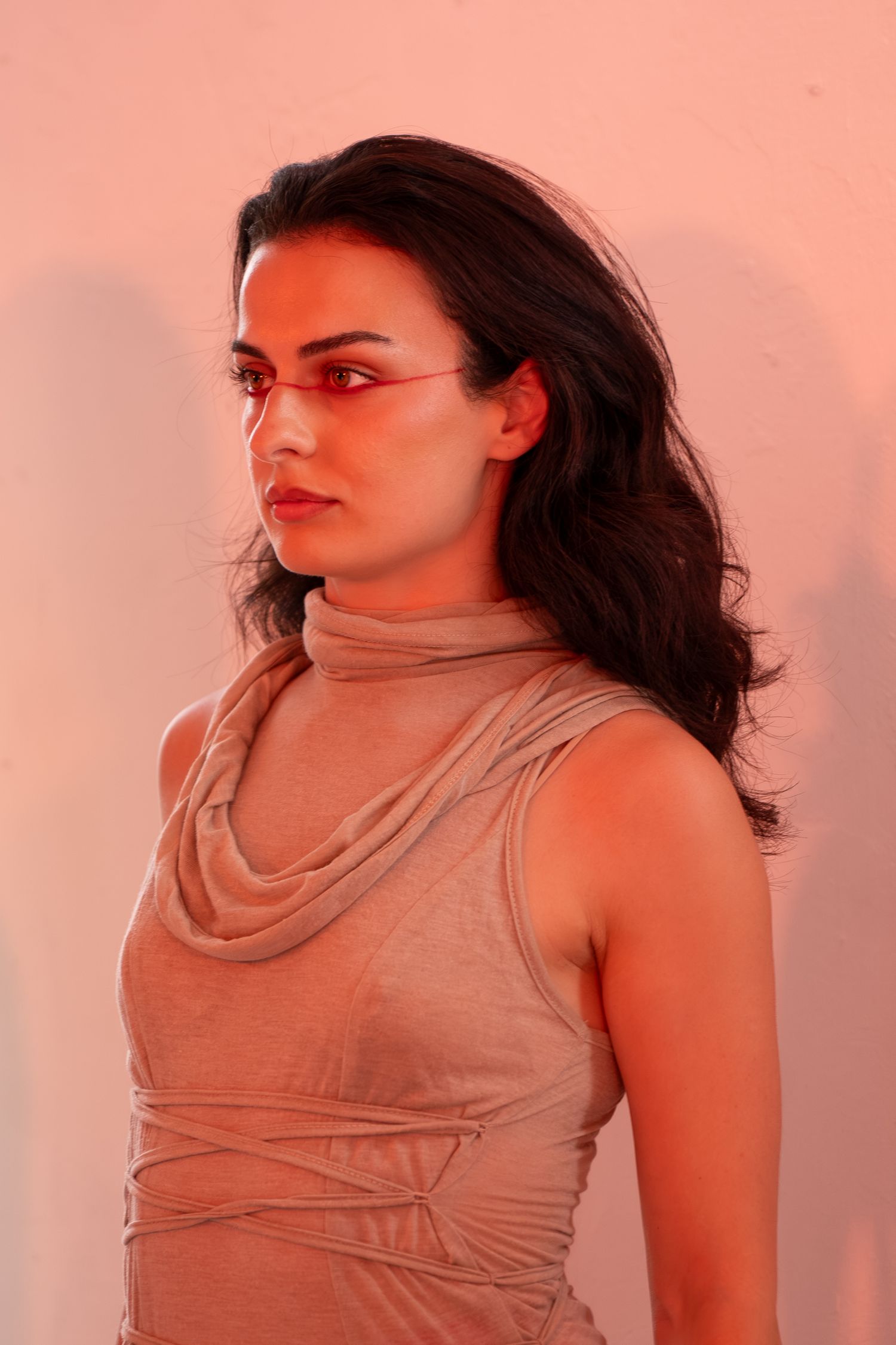 Woman with long dark wavy hair in a taupe sleeveless cowl-neck top with wrap detail, posed in profile against a warm peach backdrop.