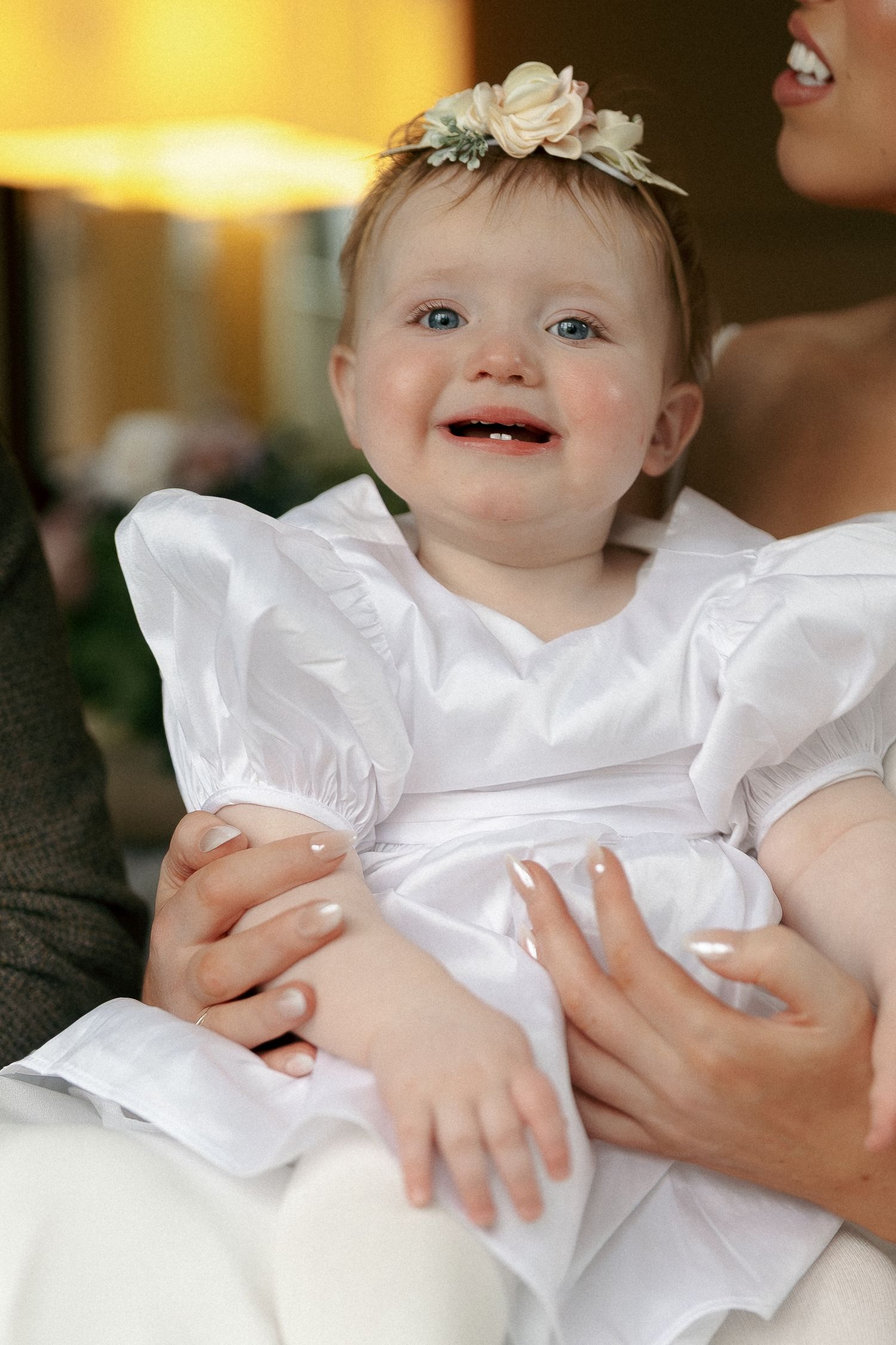 Smiling baby girl in a white dress and floral headband sits on a lap during a wedding celebration.