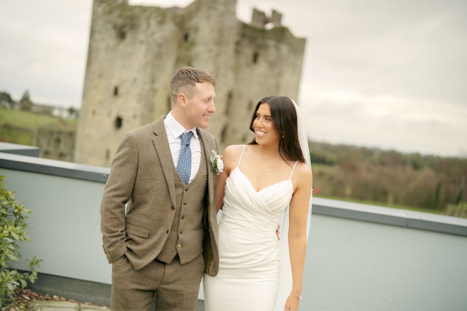 Groom in a tweed suit and bride in a white gown smile together on a terrace with a medieval castle and countryside behind.