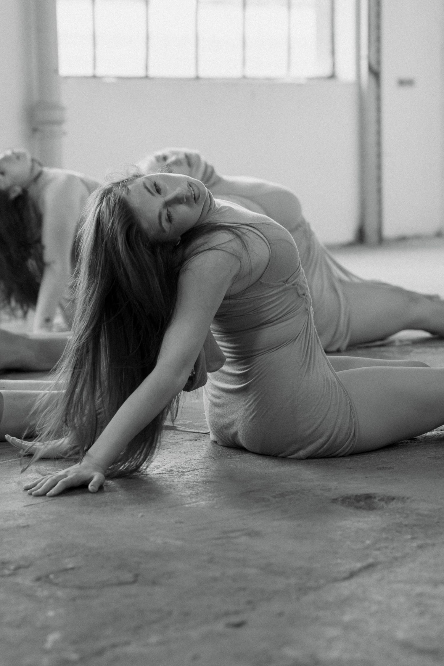 Black and white photo of dancers in a sunlit studio, one arching her back with long hair flowing to the concrete floor.