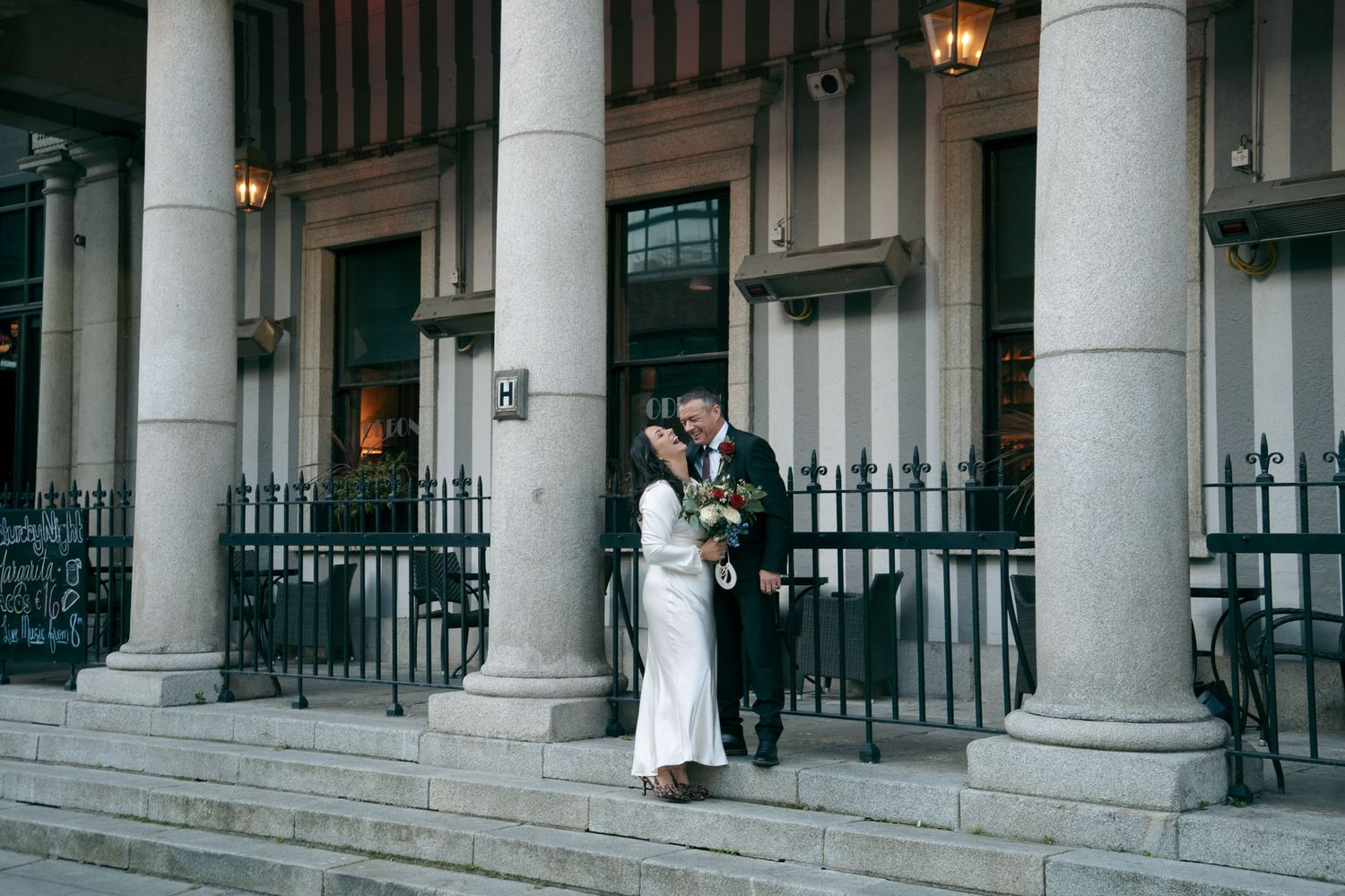 Bride and groom pose together between stone columns on the steps of a grand building with black iron railings on their wedding day.