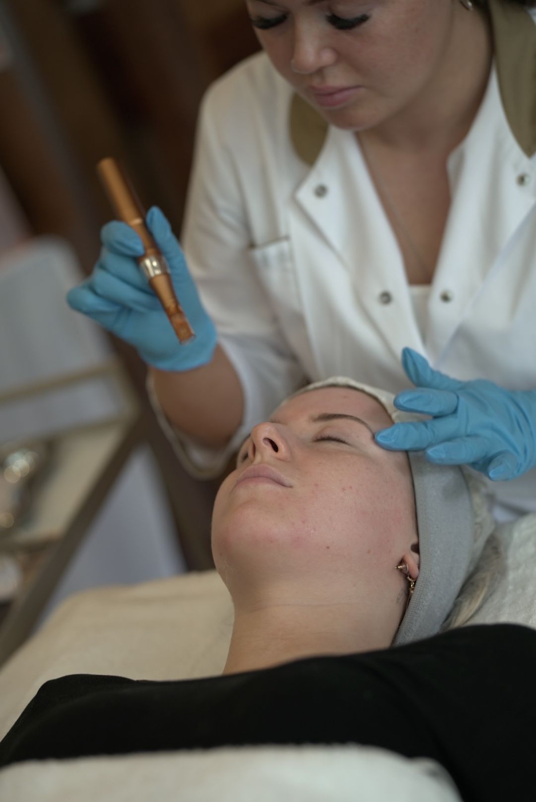 A skincare professional in a white coat and blue gloves applies a facial treatment with a copper device to a reclining client.