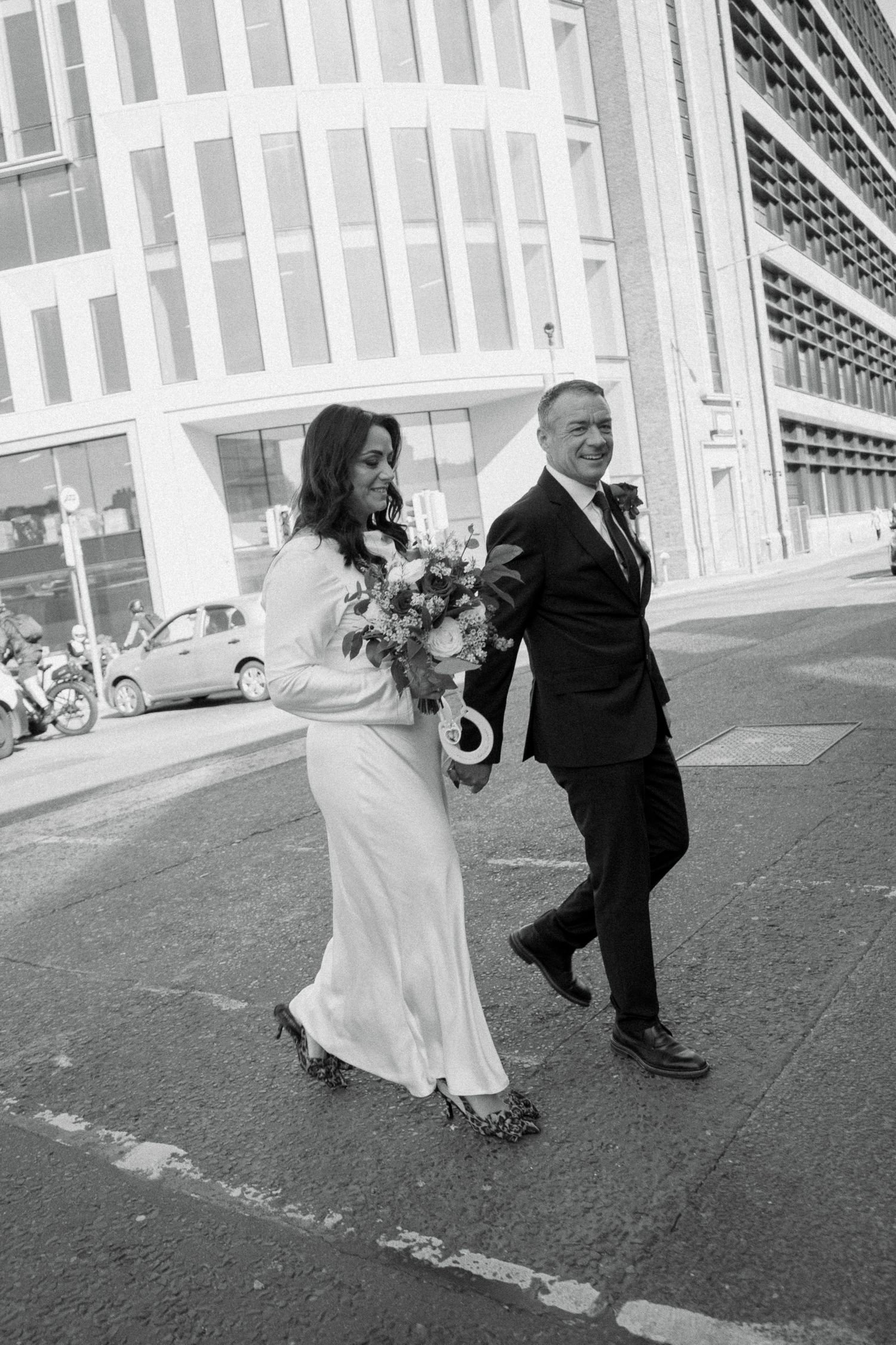 Smiling bride in a white gown holding a bouquet walks hand in hand with her groom across a city street in black and white.