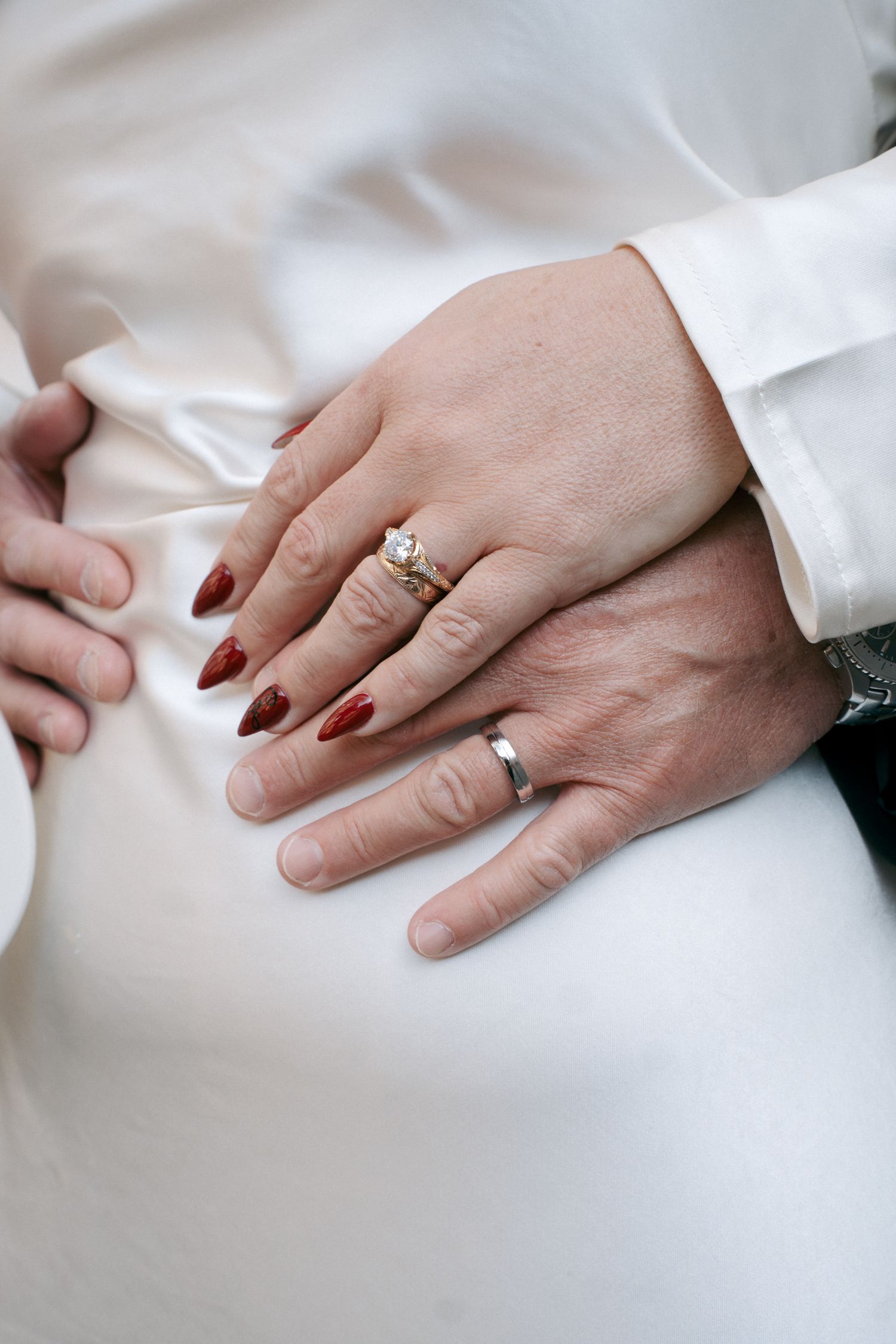 Close-up of the couple's hands resting on a white satin dress, showing a diamond engagement ring, wedding band and dark red manicured nails.