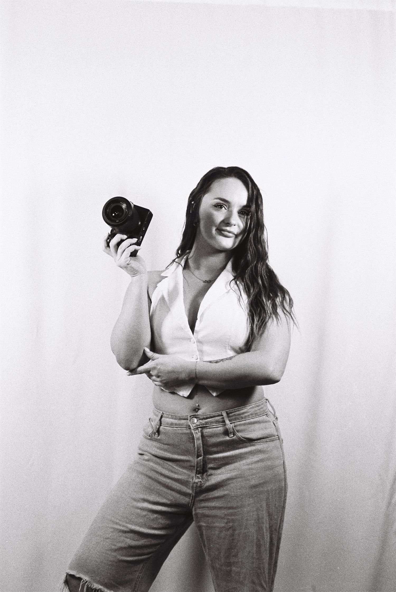 Black and white film portrait of a smiling woman in a white shirt and jeans holding a camera against a plain studio backdrop.
