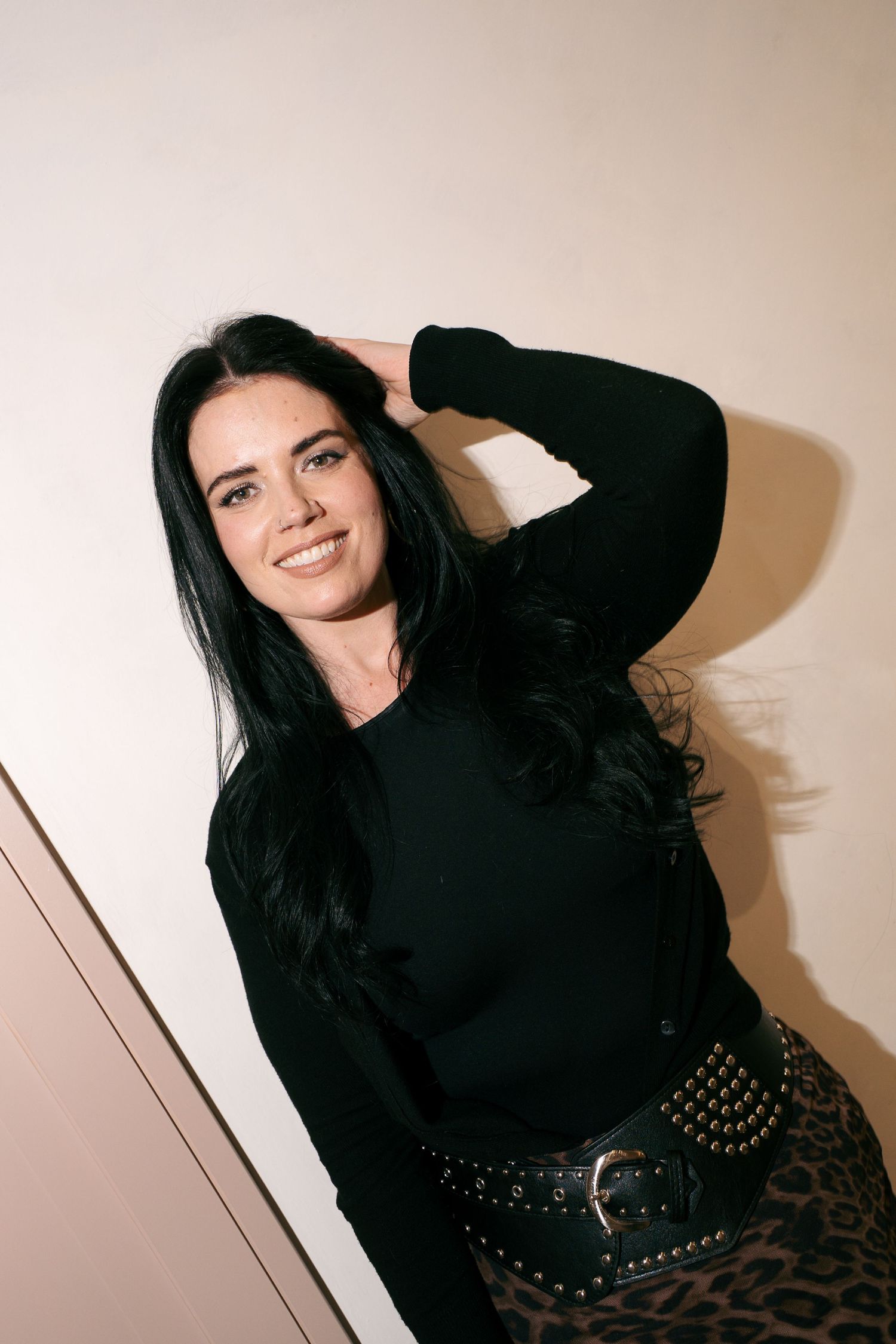 Smiling woman with long dark hair in a black top and leopard print skirt poses with a hand behind her head.