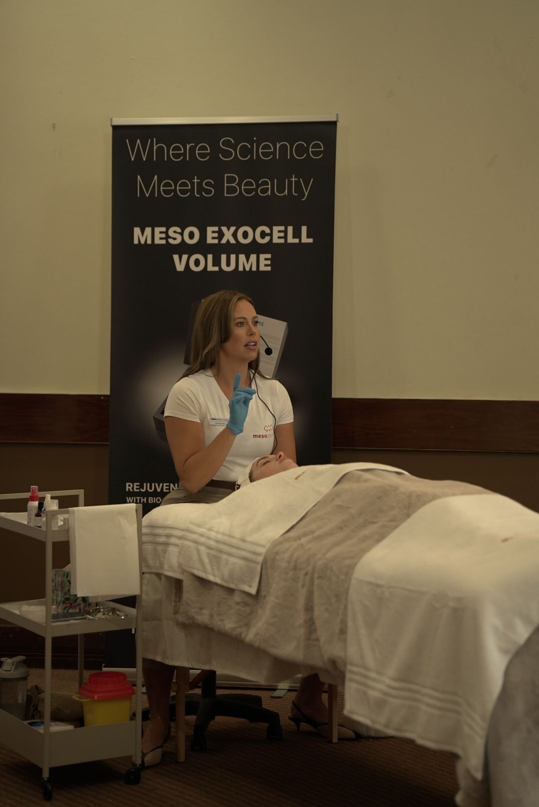 A skincare trainer wearing blue gloves speaks into a headset while demonstrating a facial treatment beside a Meso Exocell Volume banner.