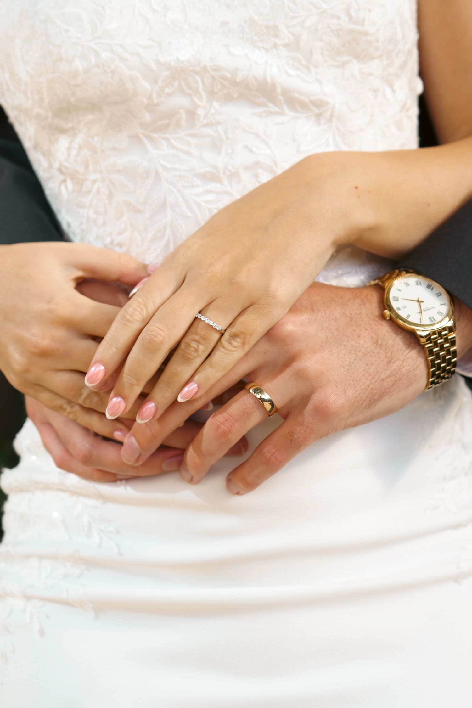 Close-up of a bride and groom's hands together showing wedding rings, pink manicured nails, and a gold wristwatch against a lace gown.