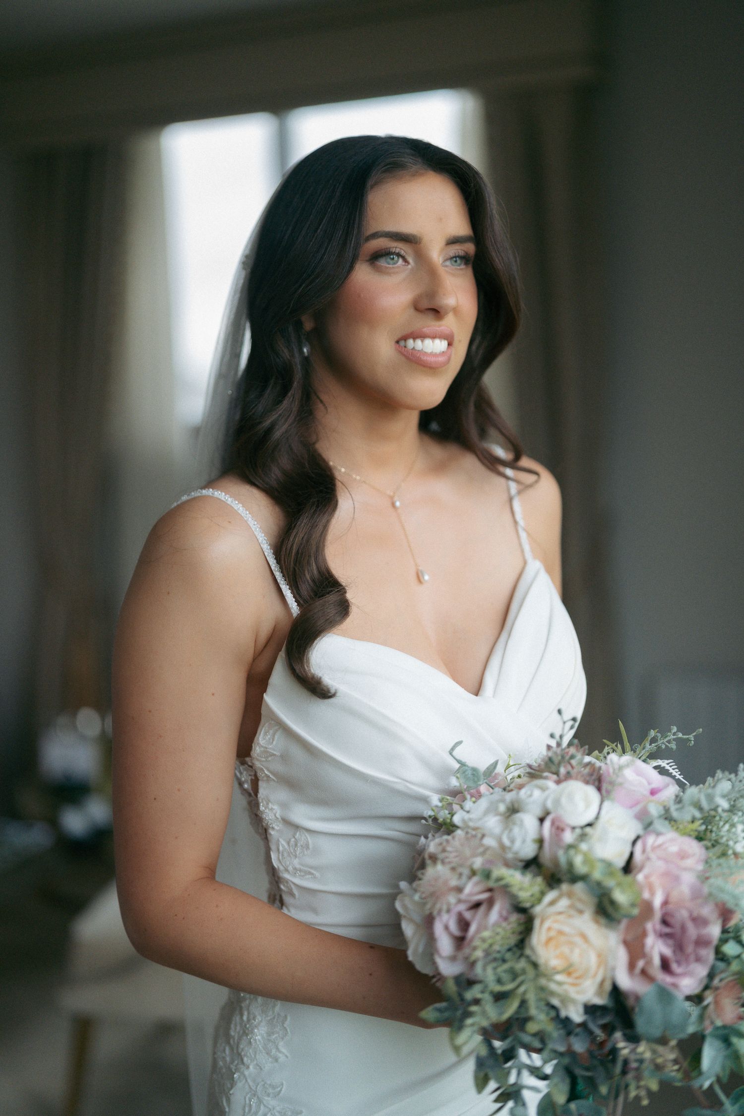 Smiling bride in a white satin wedding dress holds a soft pink and white bouquet beside a bright window.
