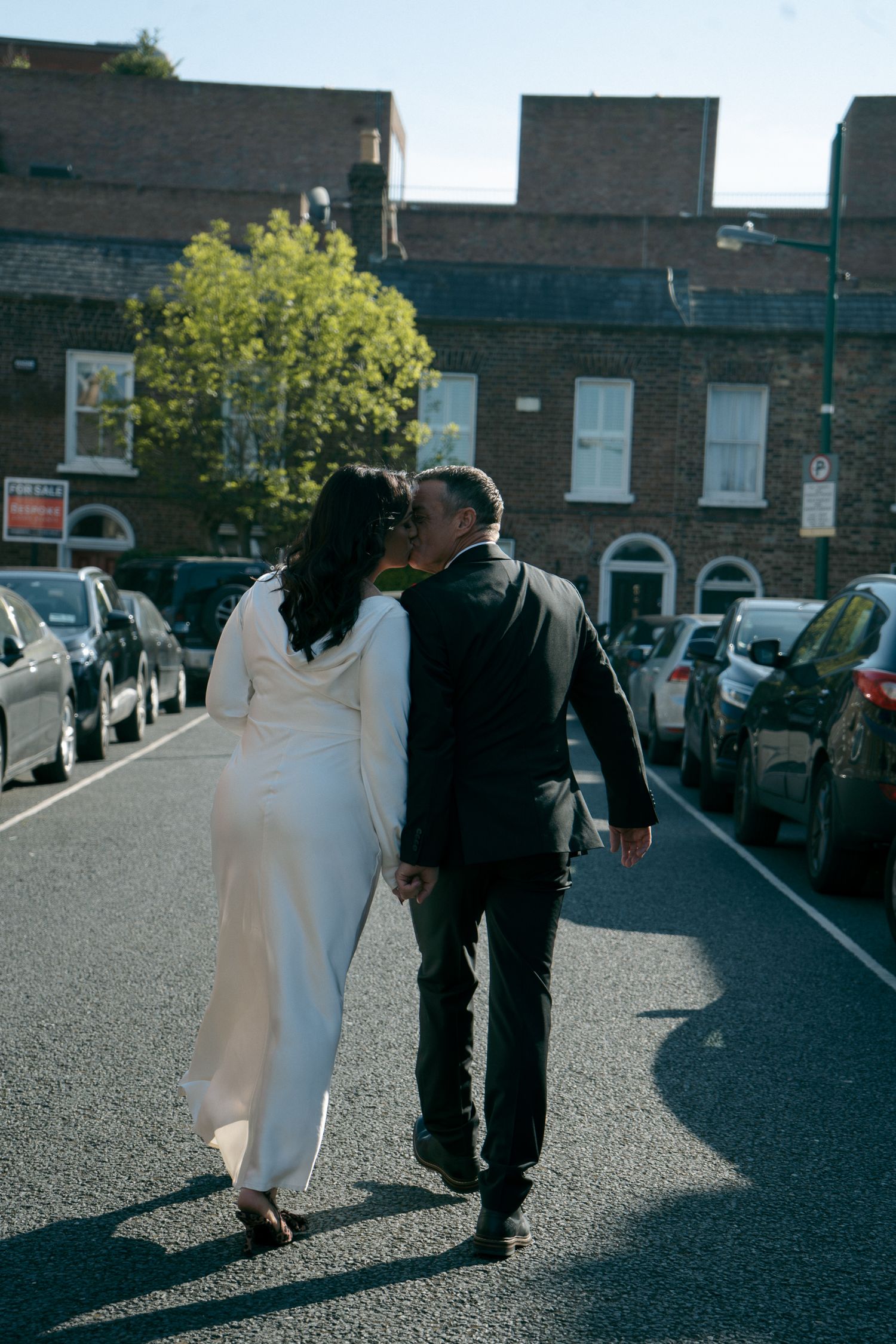 Bride in a long white dress and groom in a black suit walk arm in arm down a street lined with parked cars.